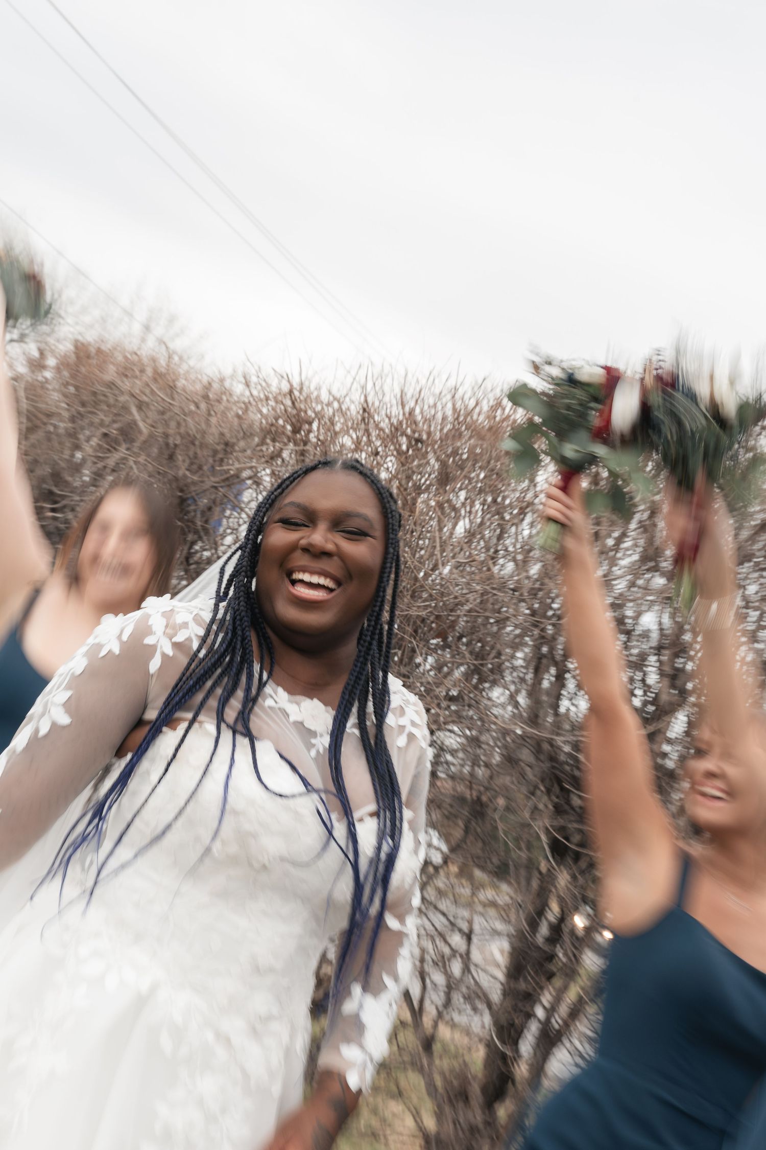 Joyful Black bride in white lace gown celebrates with bridesmaids in navy dresses, raising bouquets outdoors.