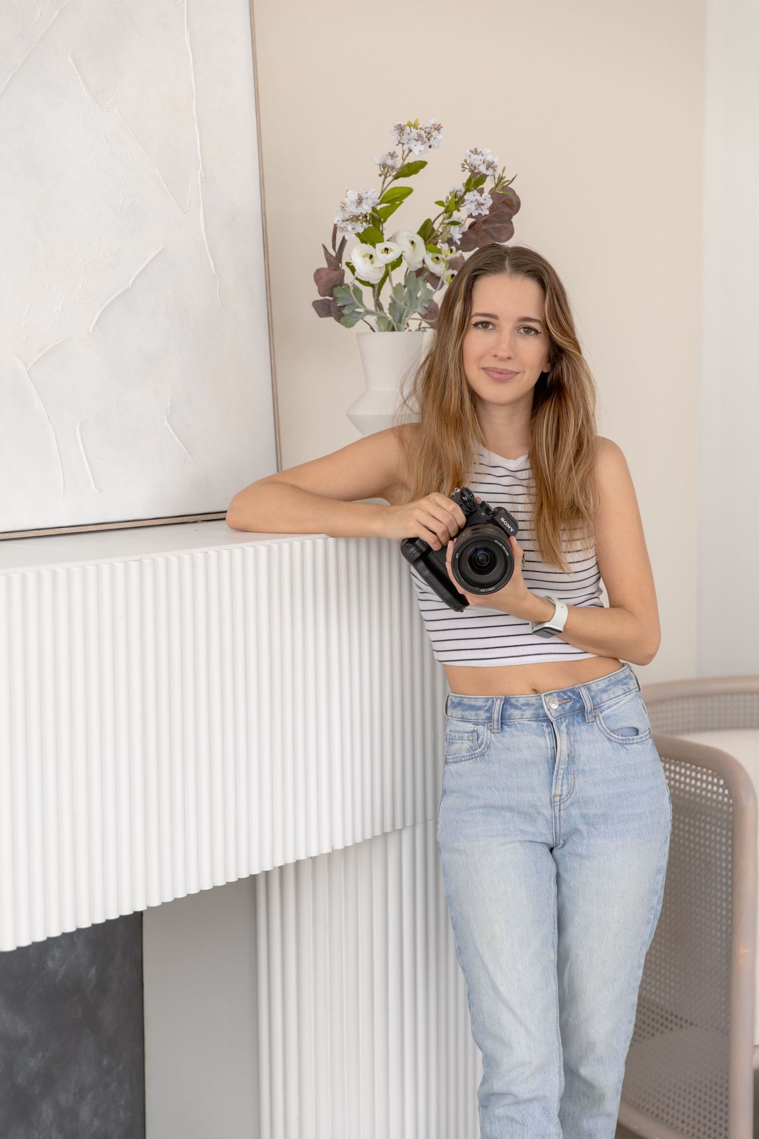 A person in a striped crop top and light wash jeans poses casually against a white wall with a camera.
