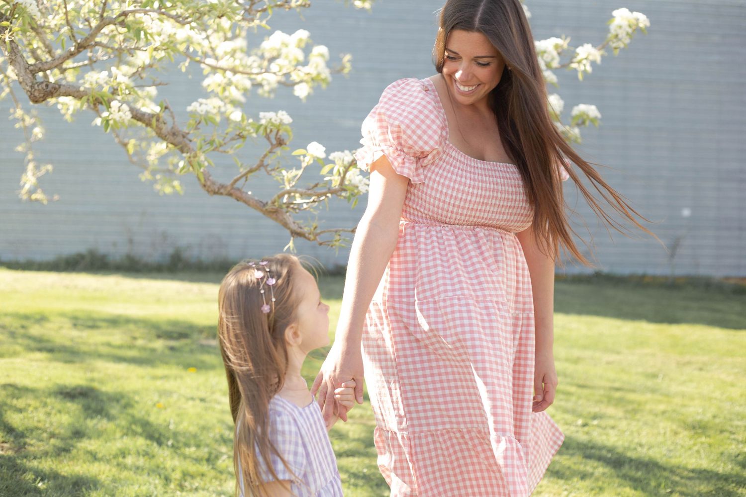 Woman in pink gingham dress poses in sunny outdoor setting with blossoming trees.