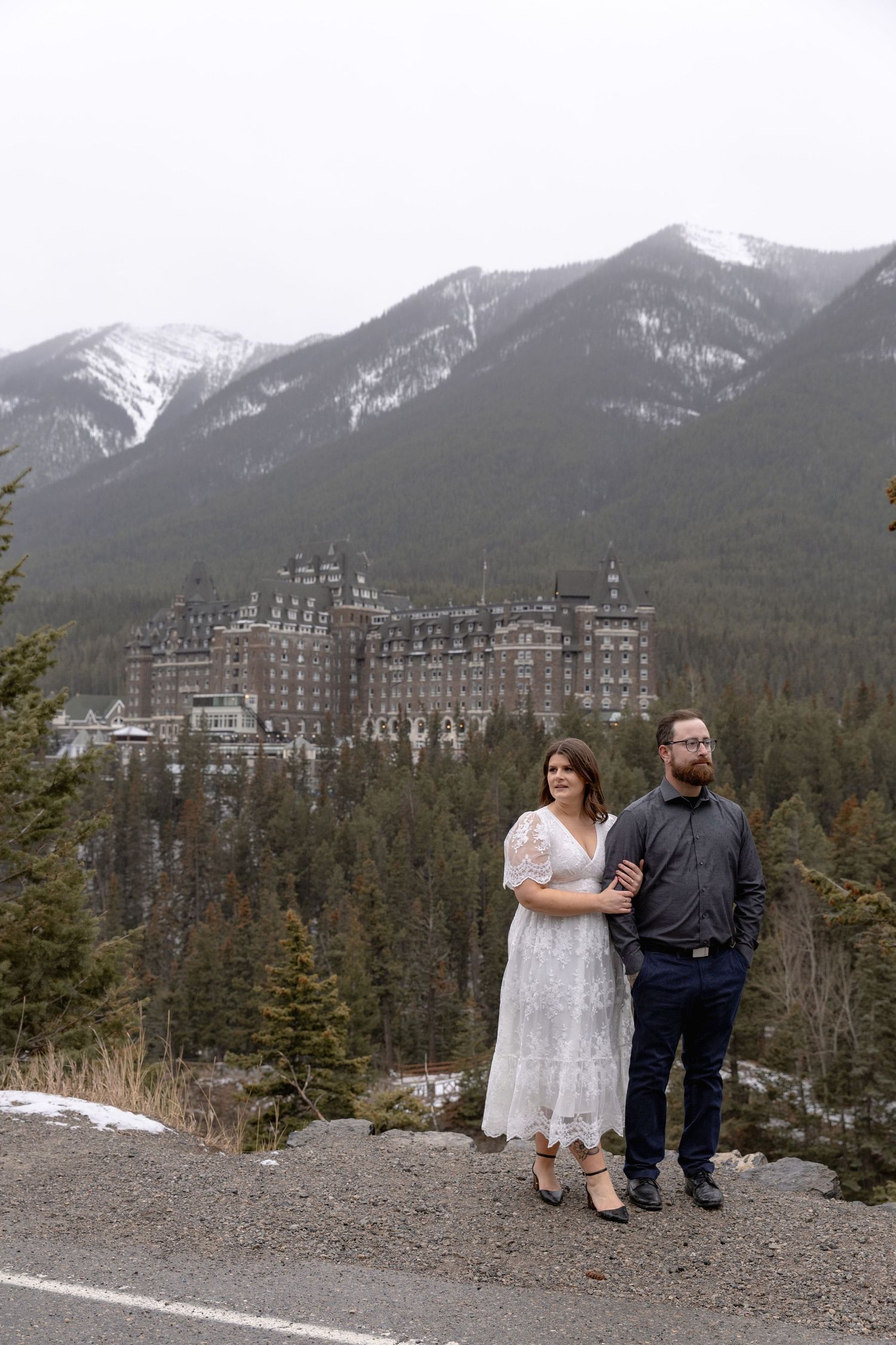A couple sits on a mountain overlook with the majestic Banff Springs Hotel and snow-capped peaks in the background.
