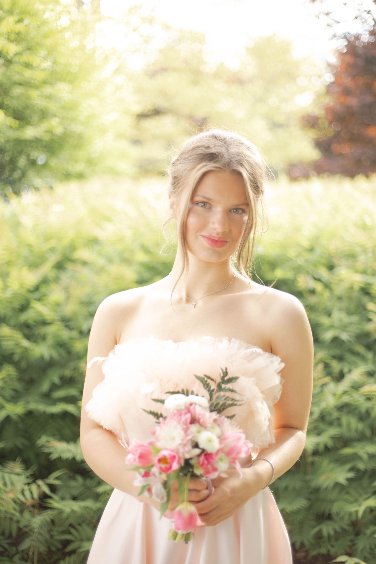A person in a blush pink dress holds a delicate bouquet of pink and white flowers against a sunlit garden backdrop.