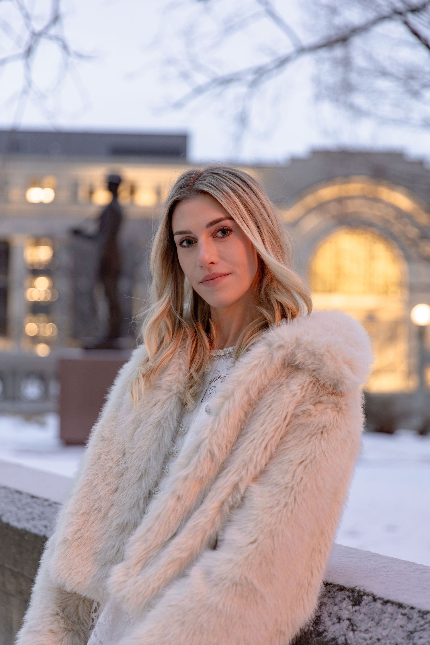 A person wearing a plush cream-colored faux fur coat poses outdoors in a snowy winter setting.