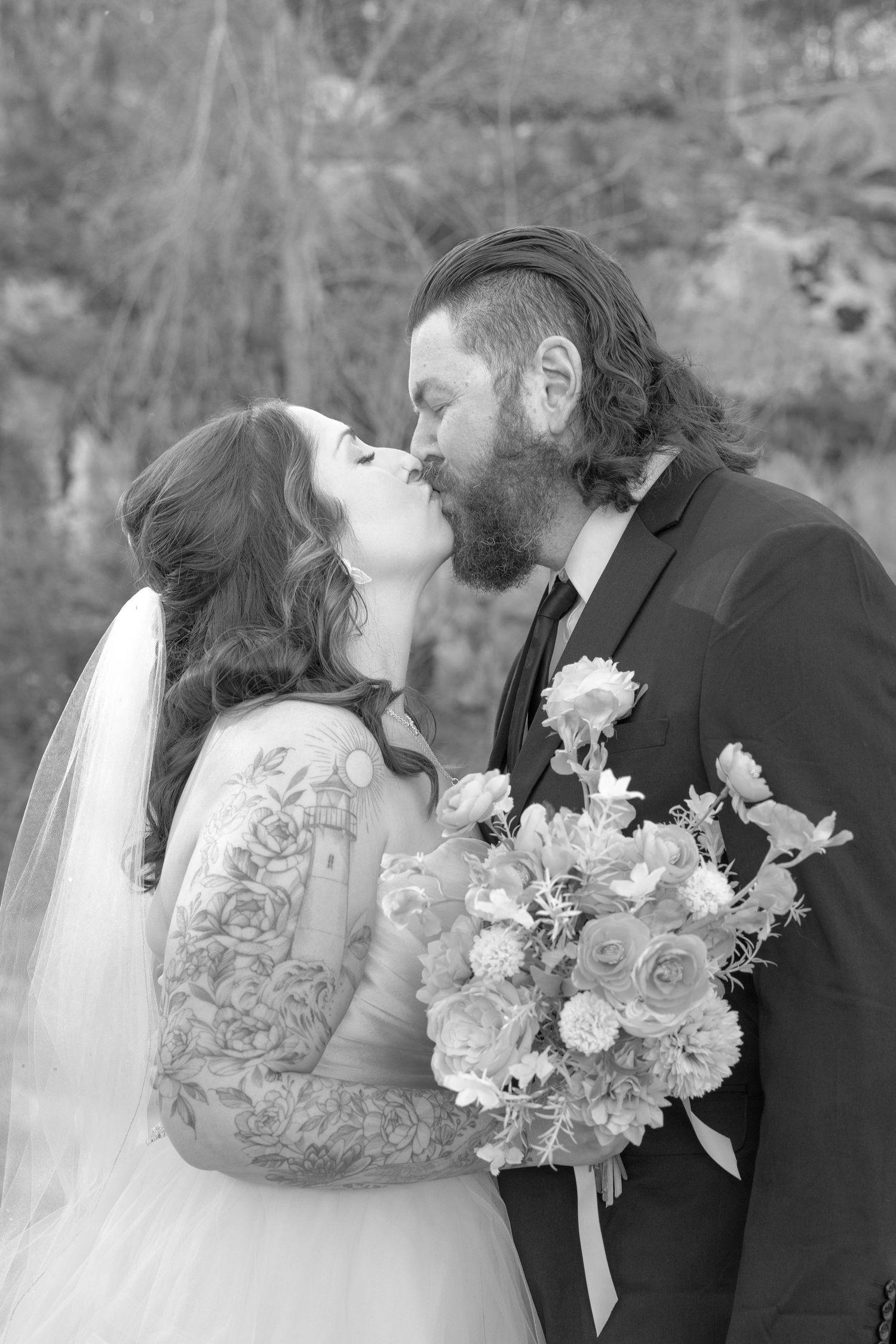 Black and white wedding portrait showing a romantic kiss between bride and groom with floral bouquet and forest backdrop.