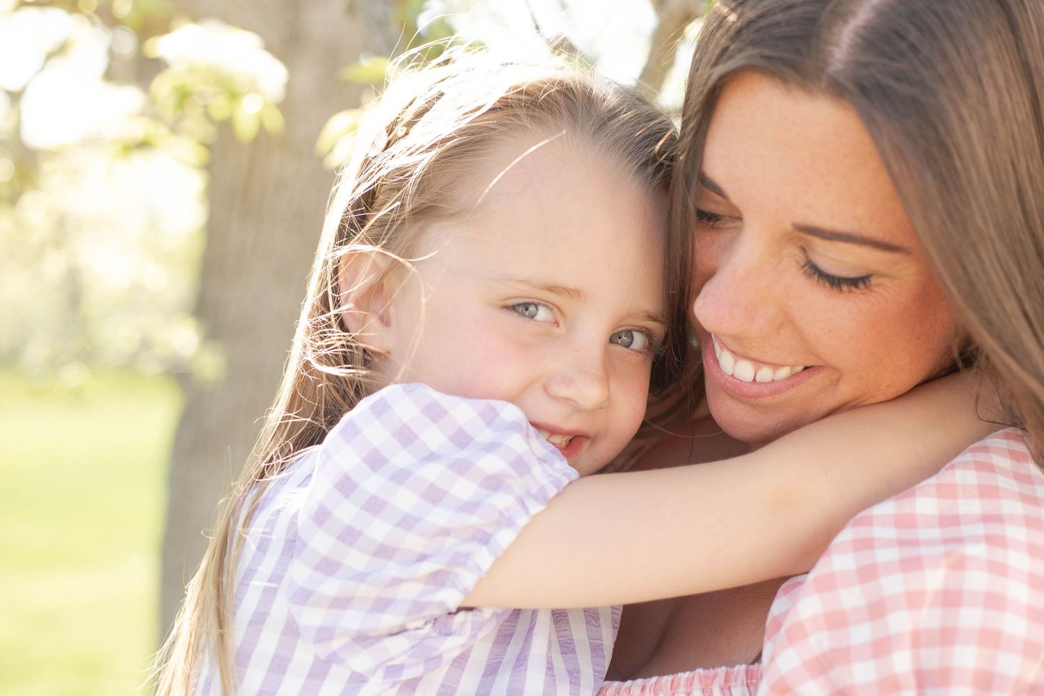 A mother and daughter wearing matching pink and white gingham share a tender embrace in warm sunlight.