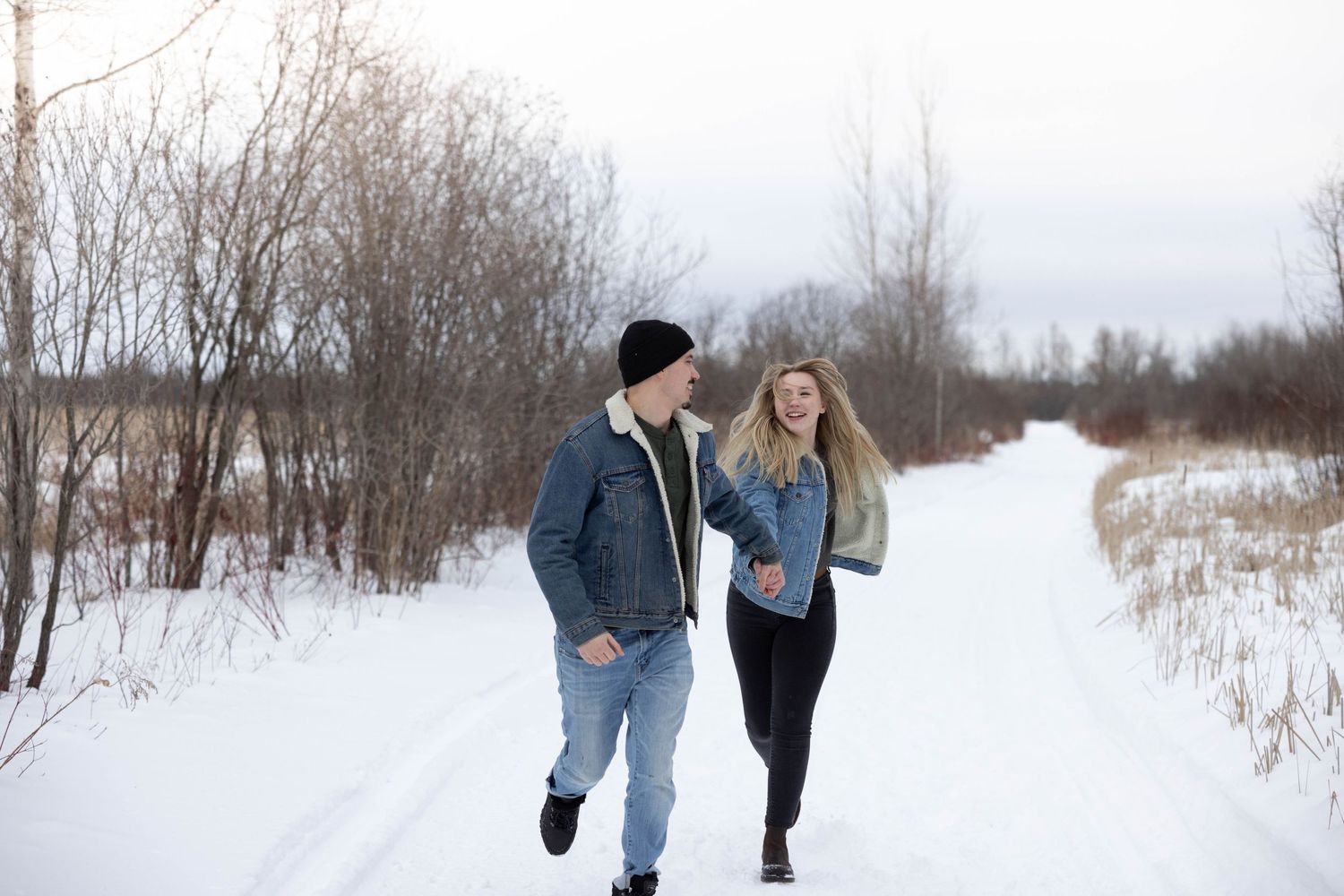 Couple walking and laughing together on a snow-covered trail surrounded by winter brush.