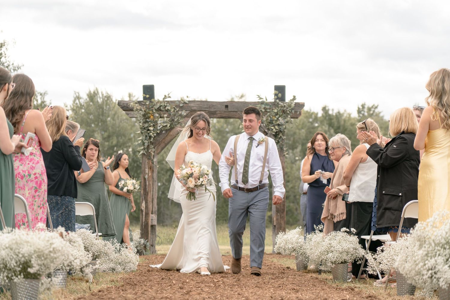 A happy couple walks down an outdoor aisle decorated with white flowers as wedding guests celebrate their union.