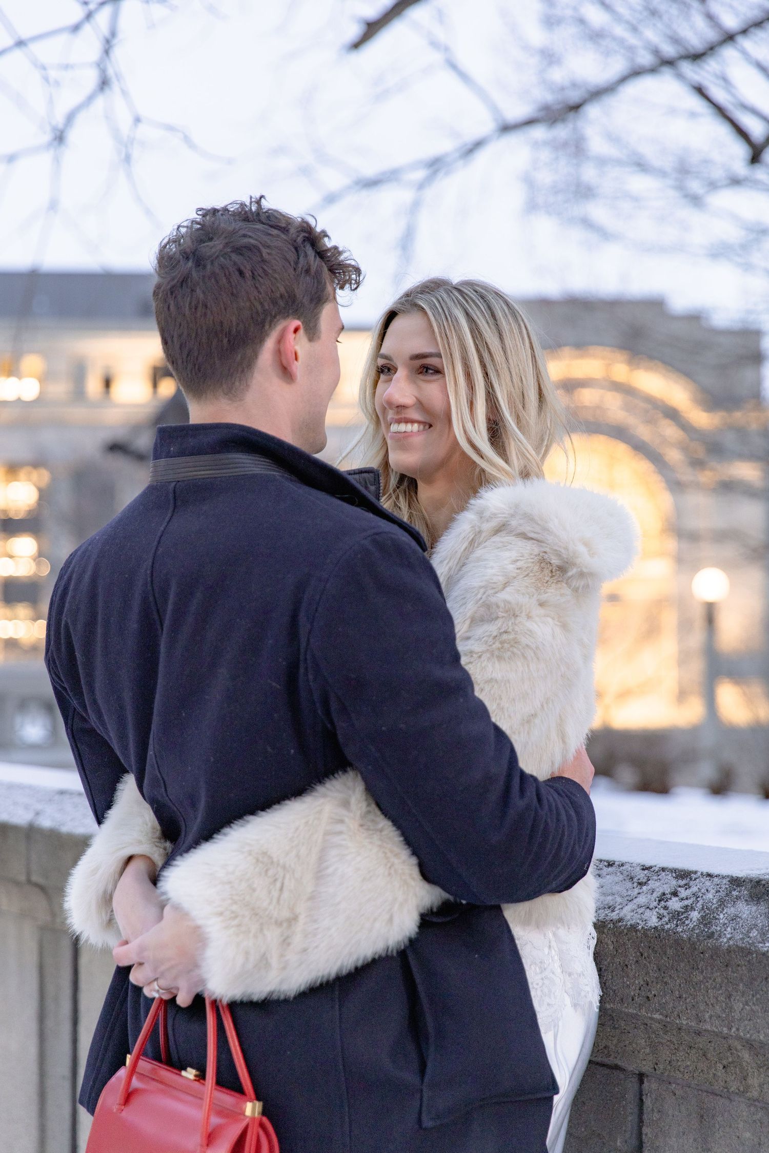 A romantic winter couple embraces outdoors near illuminated stone architecture, wearing warm coats in the evening light.