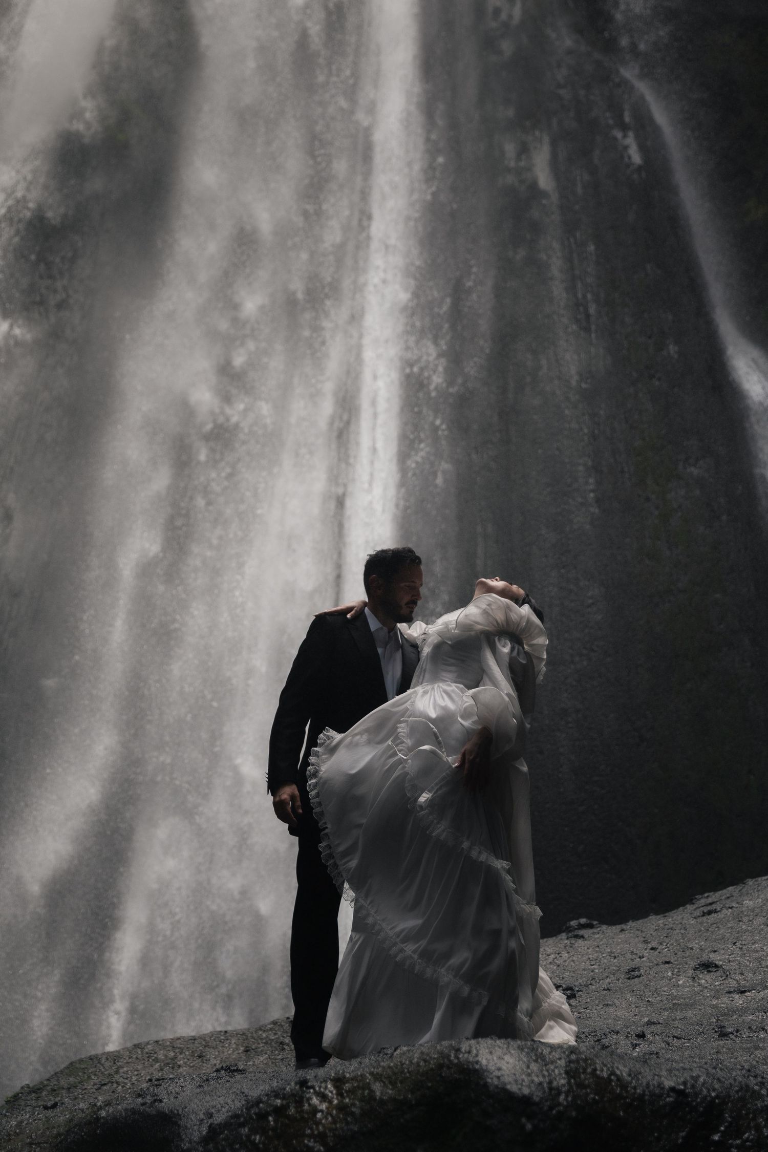 Bride and groom embrace romantically in front of a dramatic cascading waterfall on their wedding day.