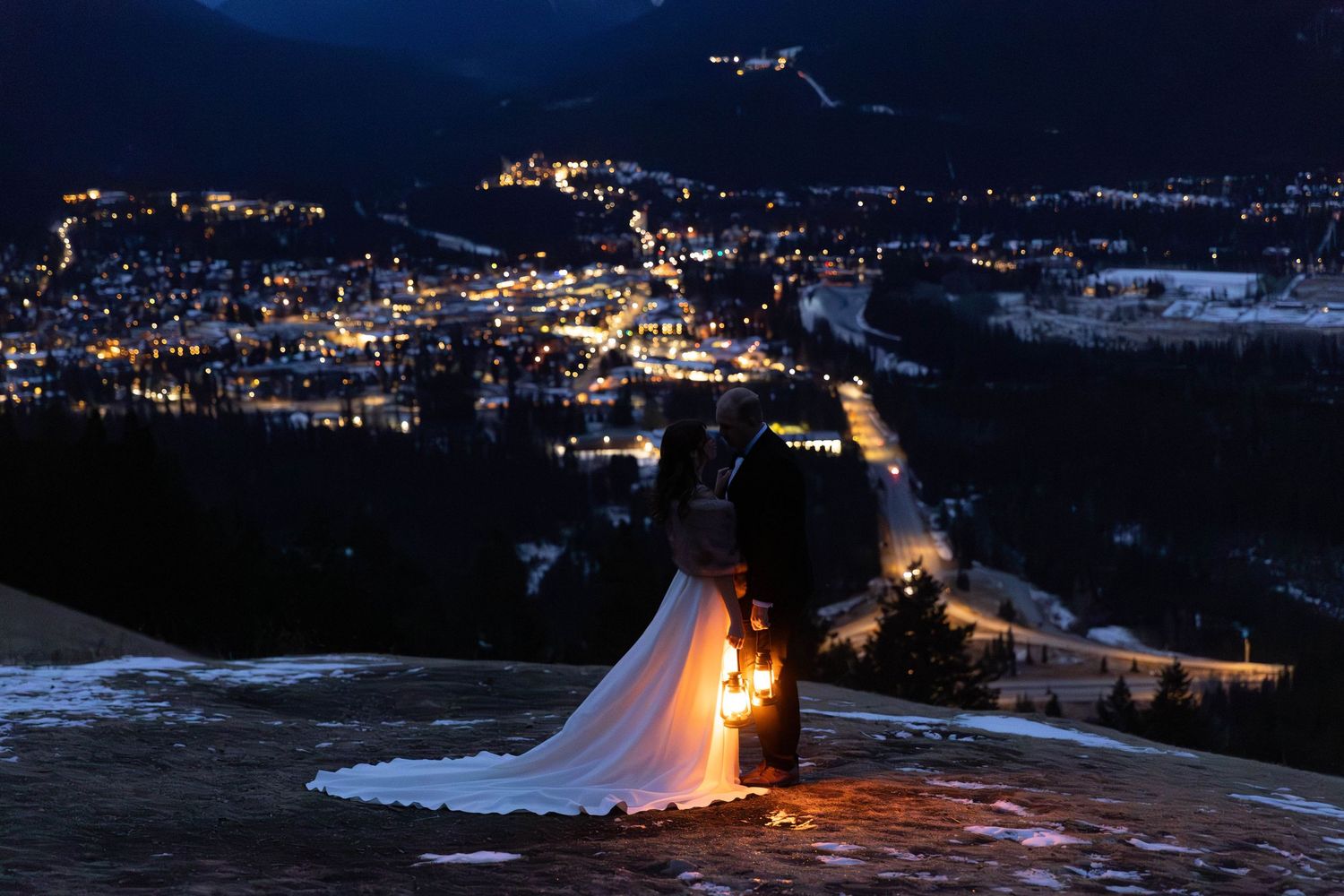 Nighttime cityscape showing illuminated buildings and streets with snow-covered mountains in the background.