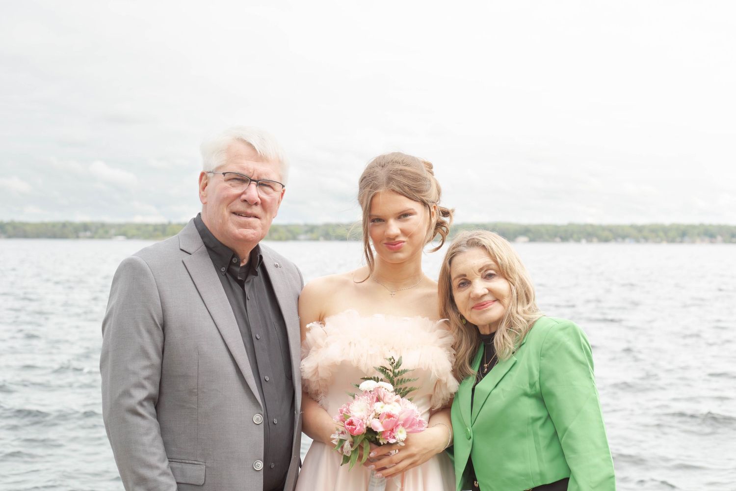 Three people pose together by a waterfront, with the person in the middle holding a pink bouquet.
