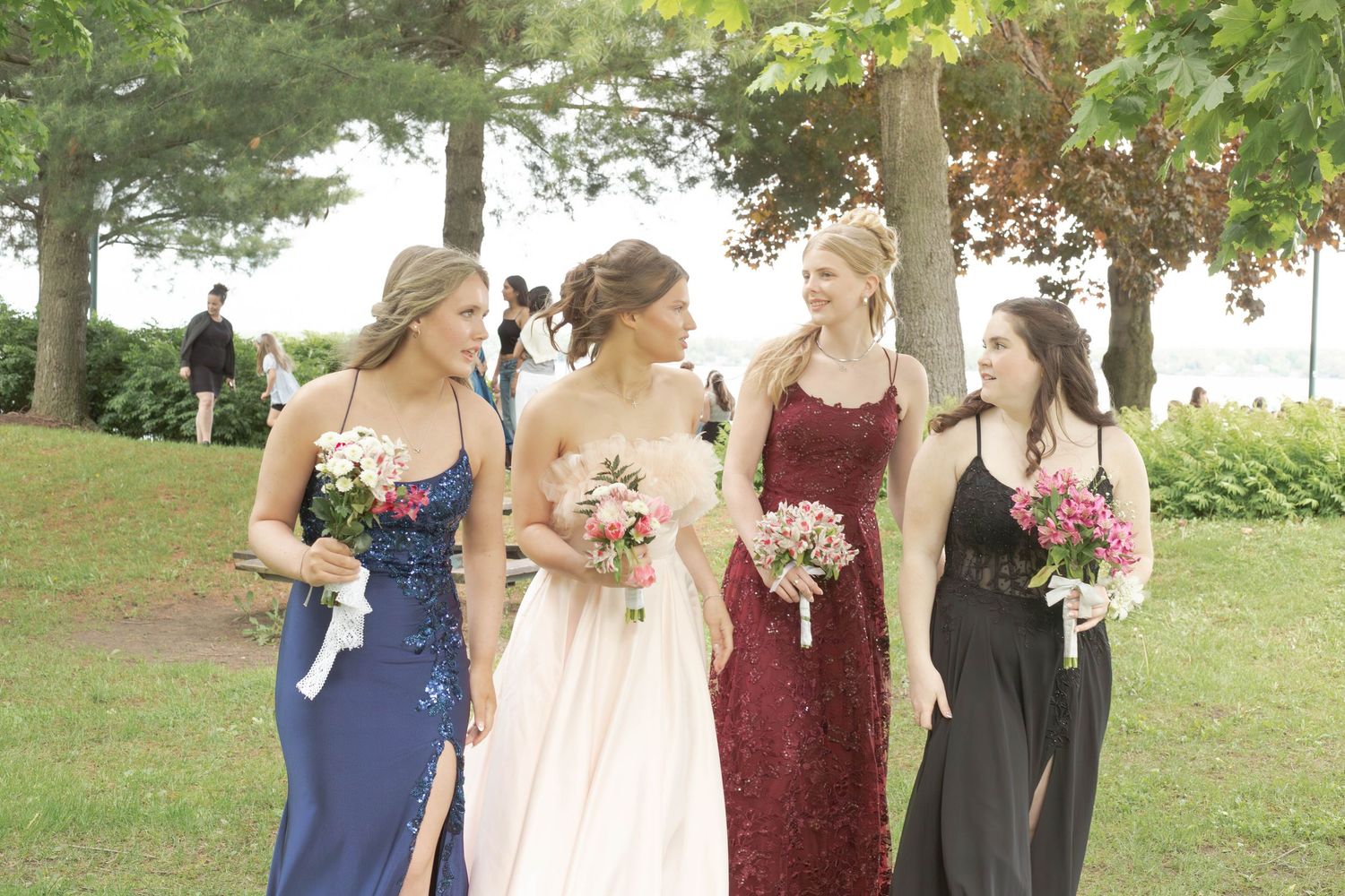 A group in formal attire stands outdoors holding pink bouquets, wearing dresses in navy, blush pink, burgundy and black.