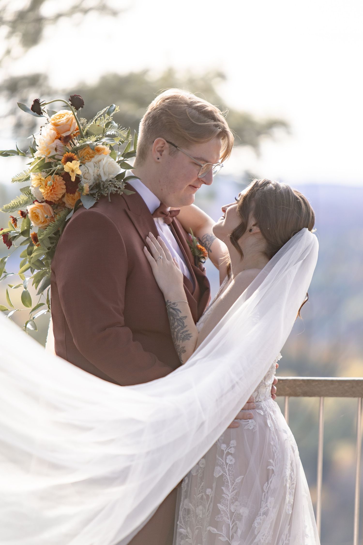 Newlywed couple embraces in romantic moment with flowing veil and peach floral bouquet against scenic mountain backdrop.