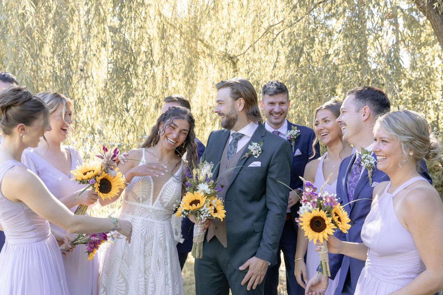 Wedding party in purple dresses and black suits pose outdoors with bright yellow sunflower bouquets.