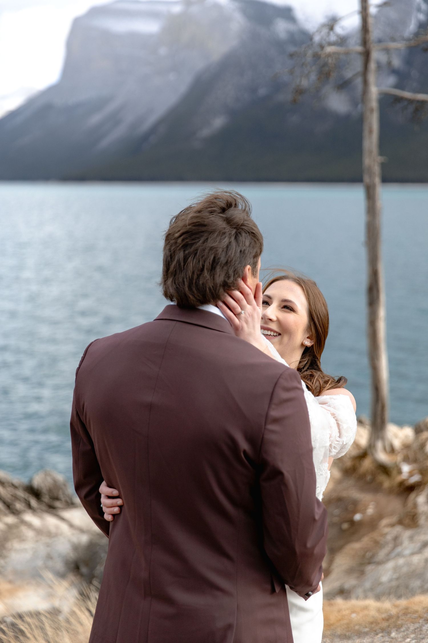 A couple shares an intimate moment by a scenic mountain lake with snow-capped peaks in the background.