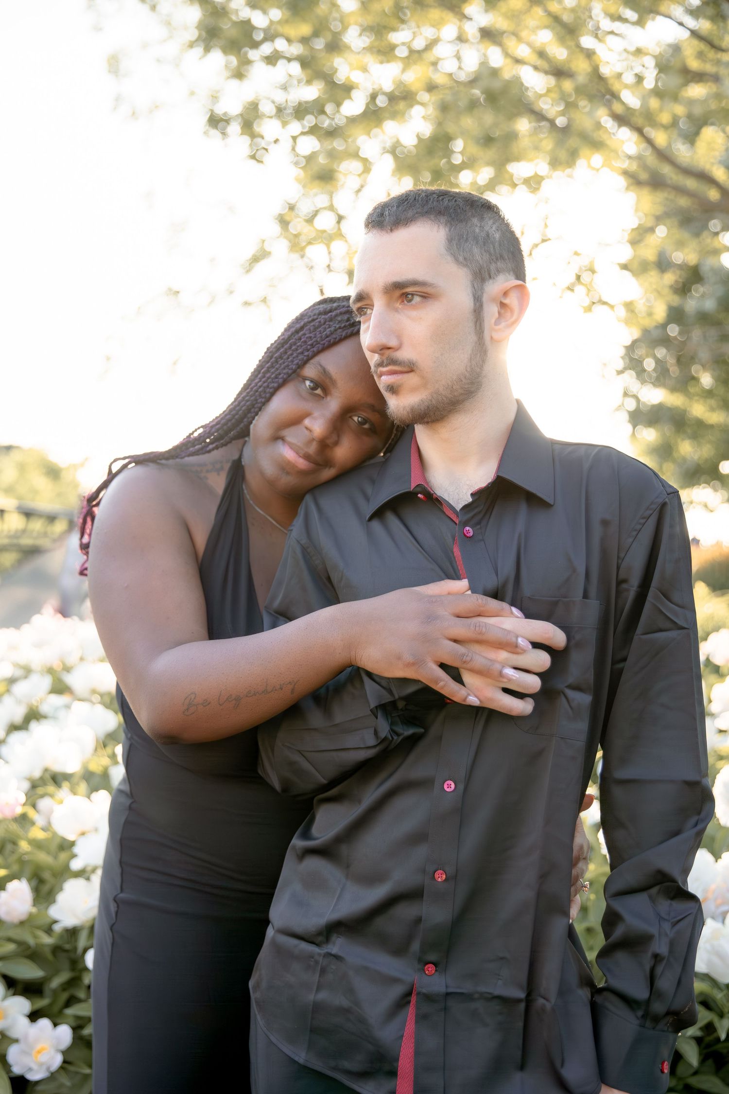 A romantic couple in black clothing embraces in a sunlit outdoor setting with trees in the background.