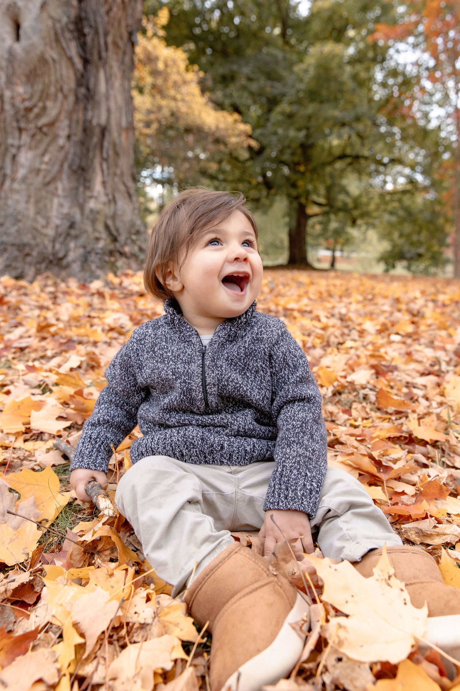A child plays joyfully in a pile of fallen autumn leaves while wearing a cozy gray sweater.