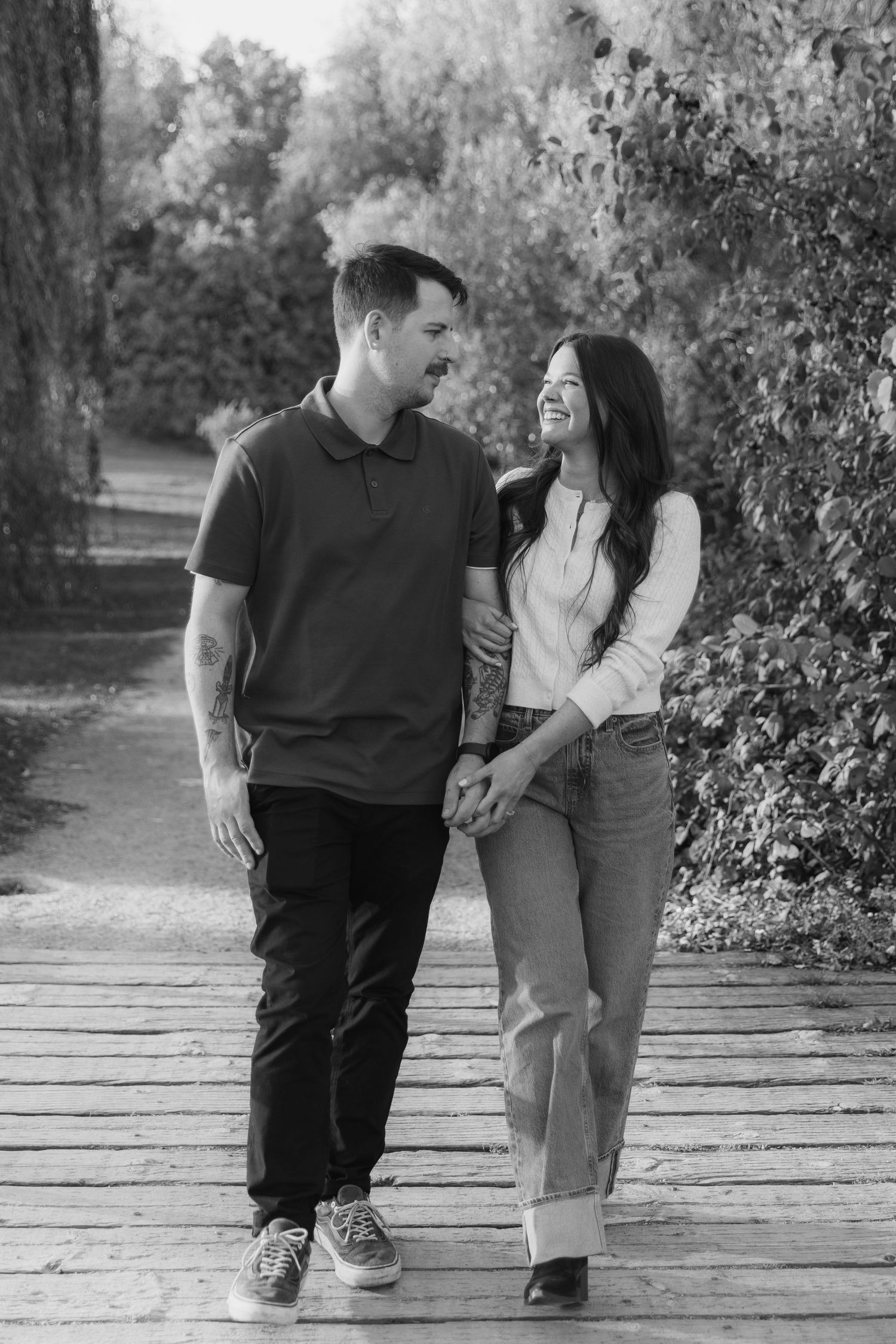 A couple walks together on a wooden bridge path through an autumn park setting.