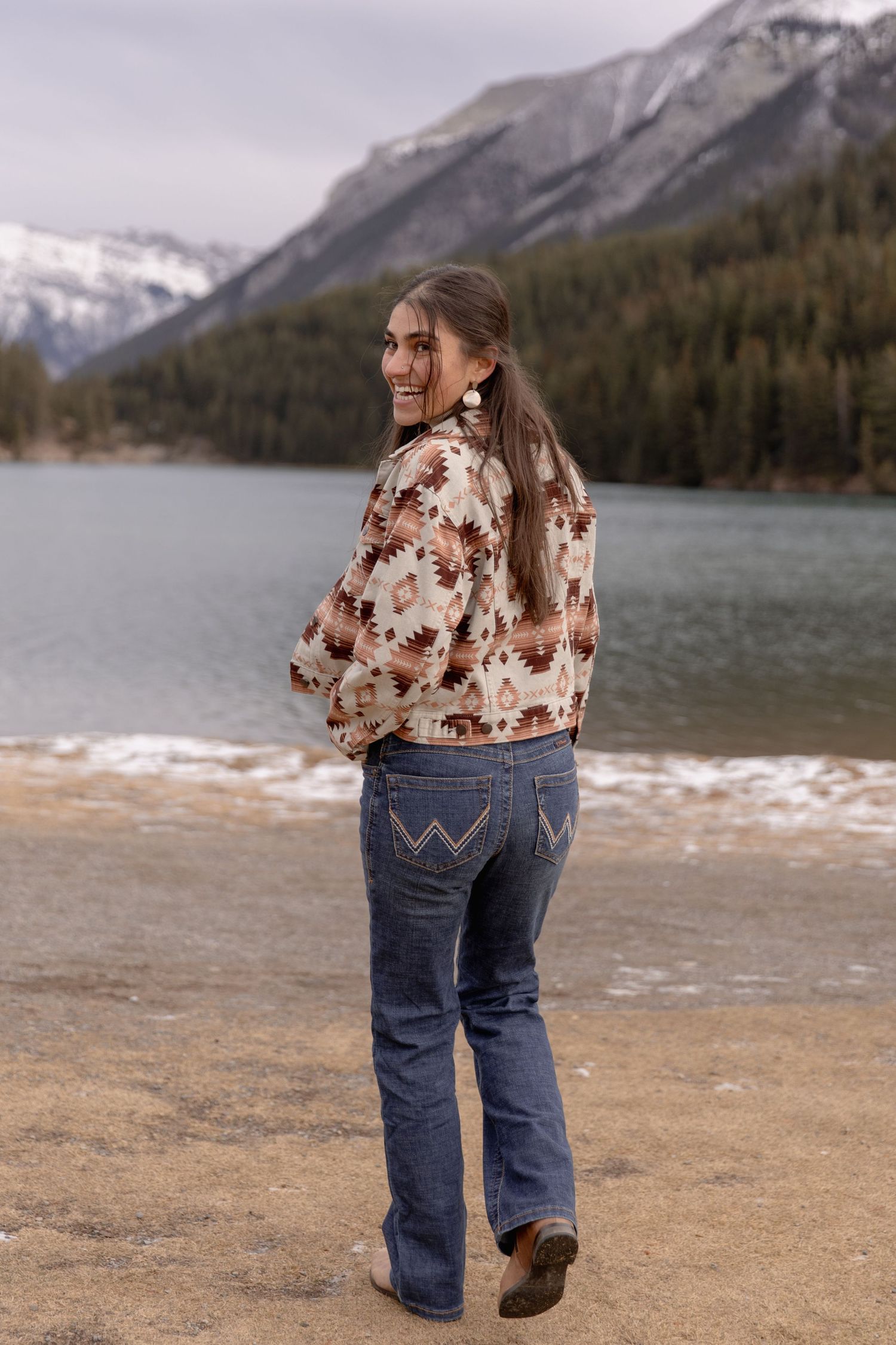 Woman in southwestern print jacket walks along shoreline with snow-capped mountains in background.