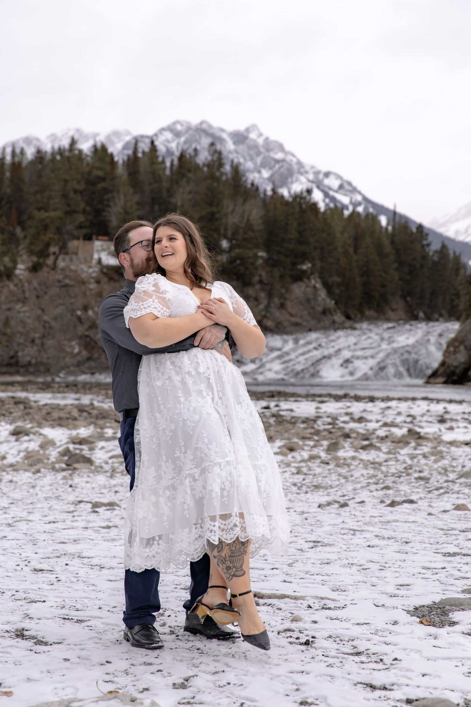 A couple embraces in a snowy mountain landscape with evergreen trees and peaks in the background.