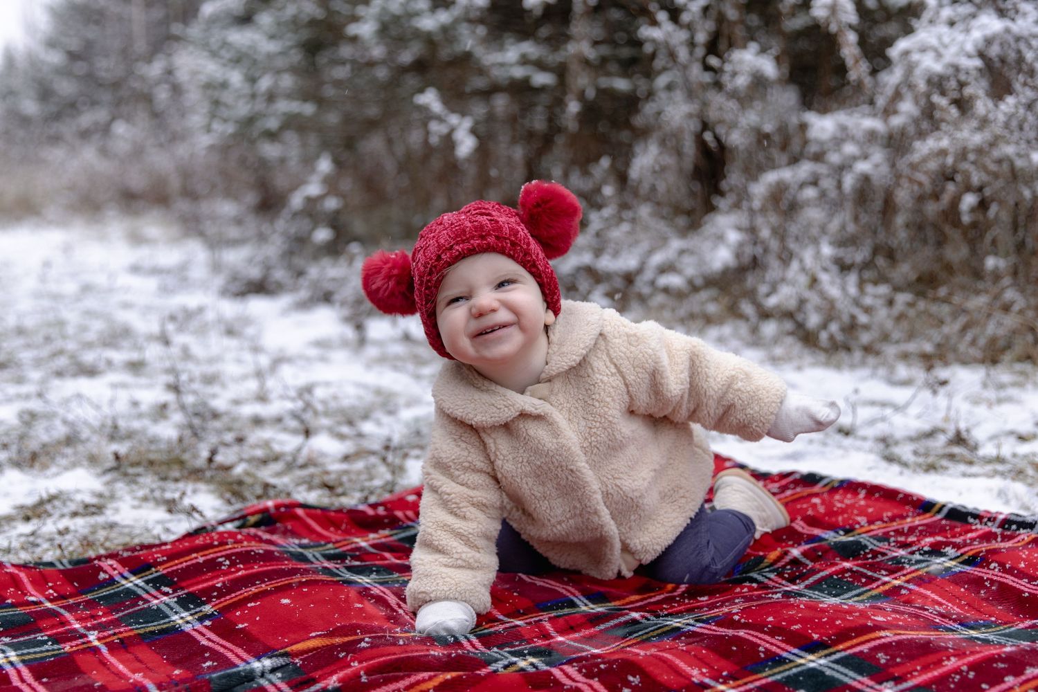 A small child sits on a red plaid blanket in the snow wearing a cozy beige sweater and red pom-pom hat.