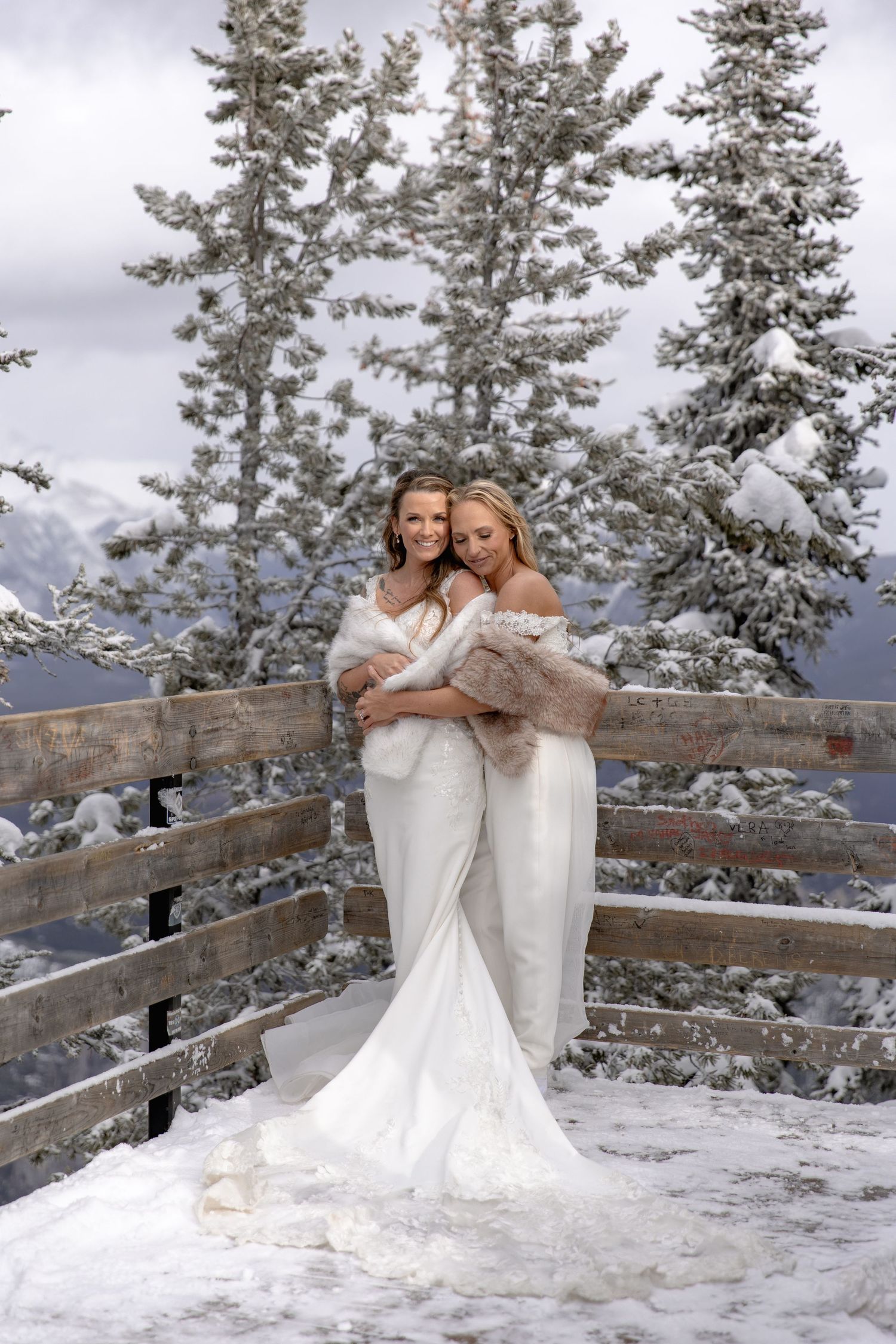 Two people in white gowns pose together against a snowy mountain backdrop with evergreen trees.