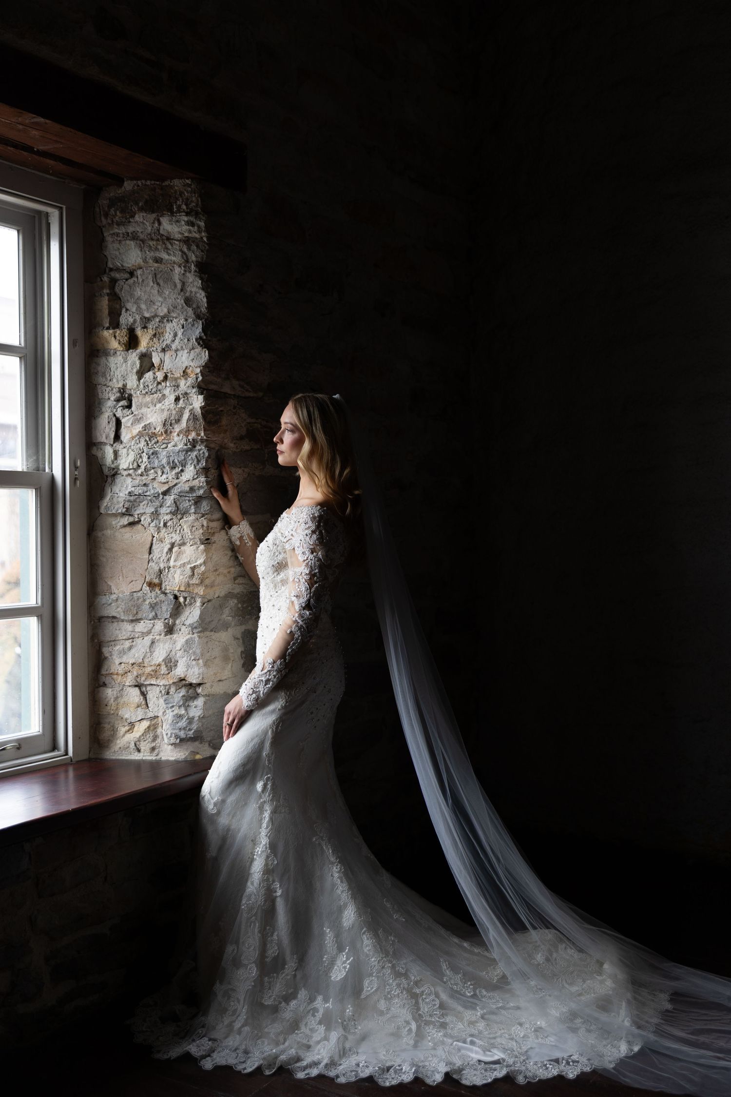A bride in a lace wedding gown with flowing veil poses by a rustic stone wall near a window in dramatic lighting.