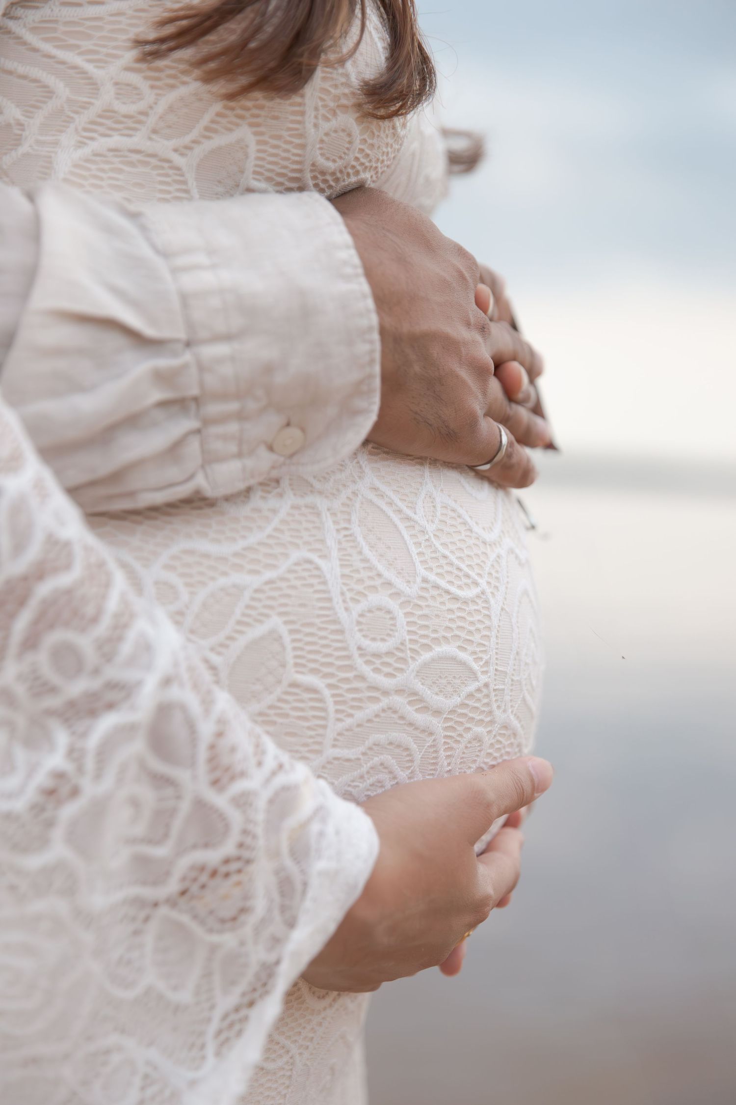Close-up of pregnant woman's belly in white lace dress with hands cradling her bump against soft background.