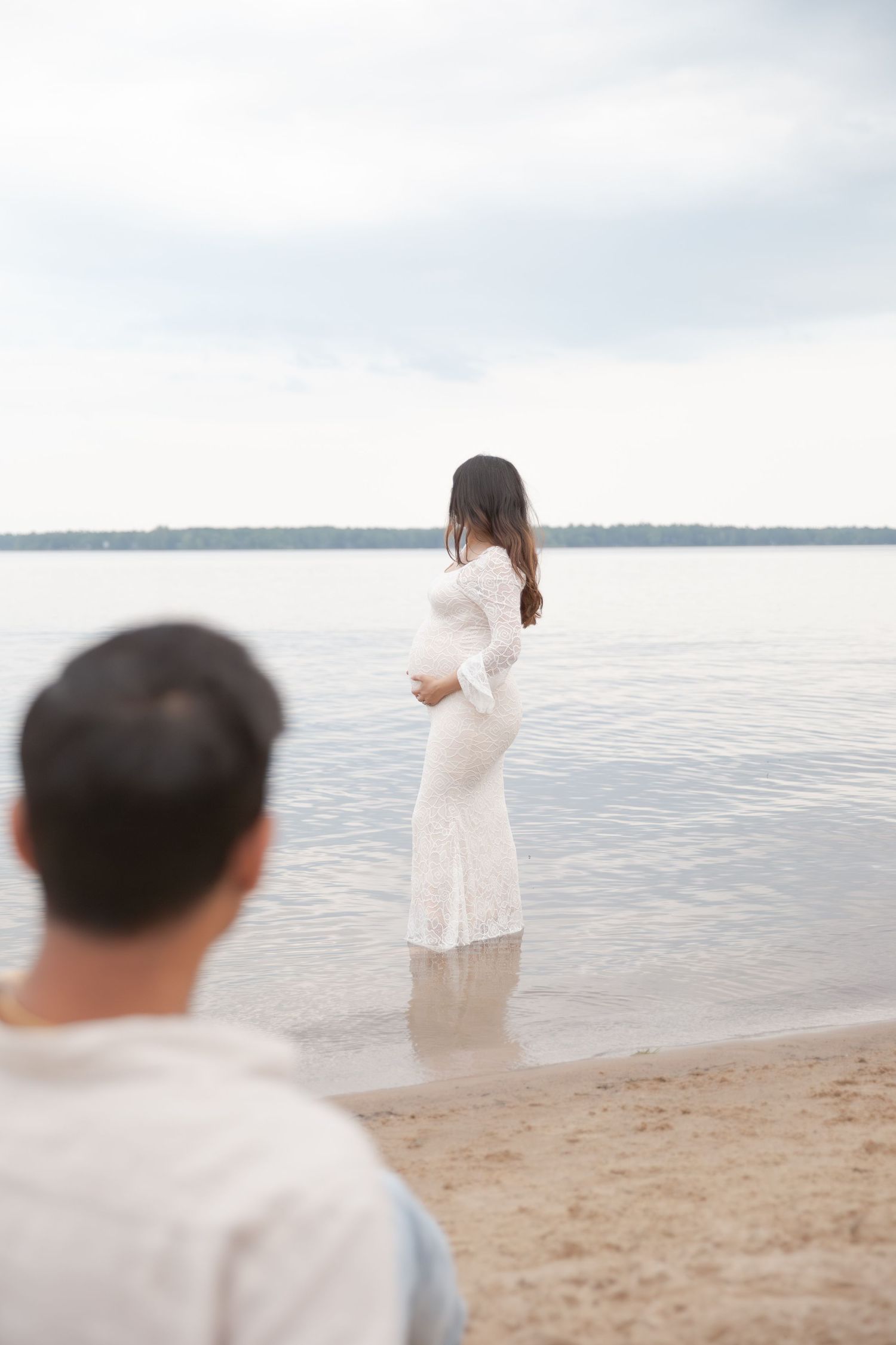 A figure in flowing white clothing walks along a serene beach shoreline on a misty overcast day.