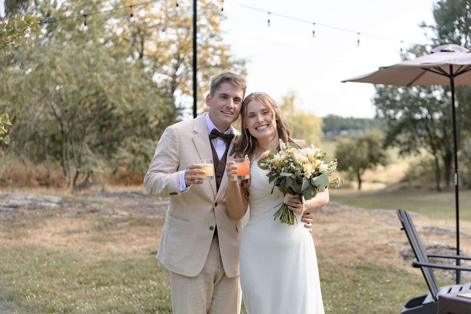 A couple in light-colored wedding attire poses together outdoors on a sunny autumn day in a natural setting.