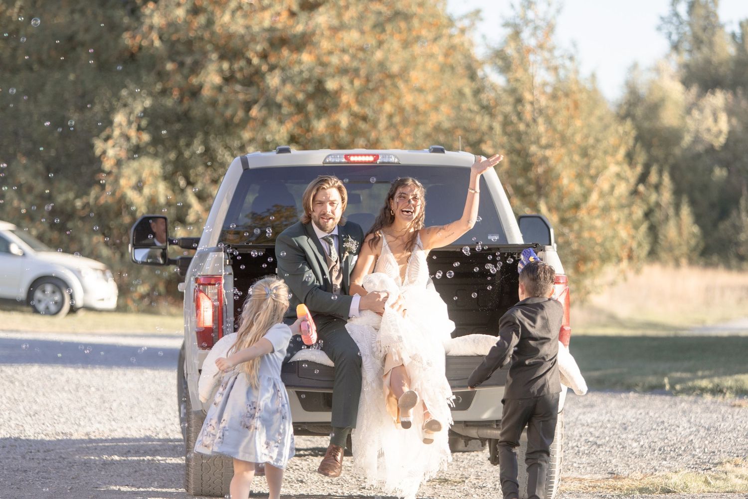 A wedding party walks together on a gravel path while celebrating and laughing outdoors on a sunny day.