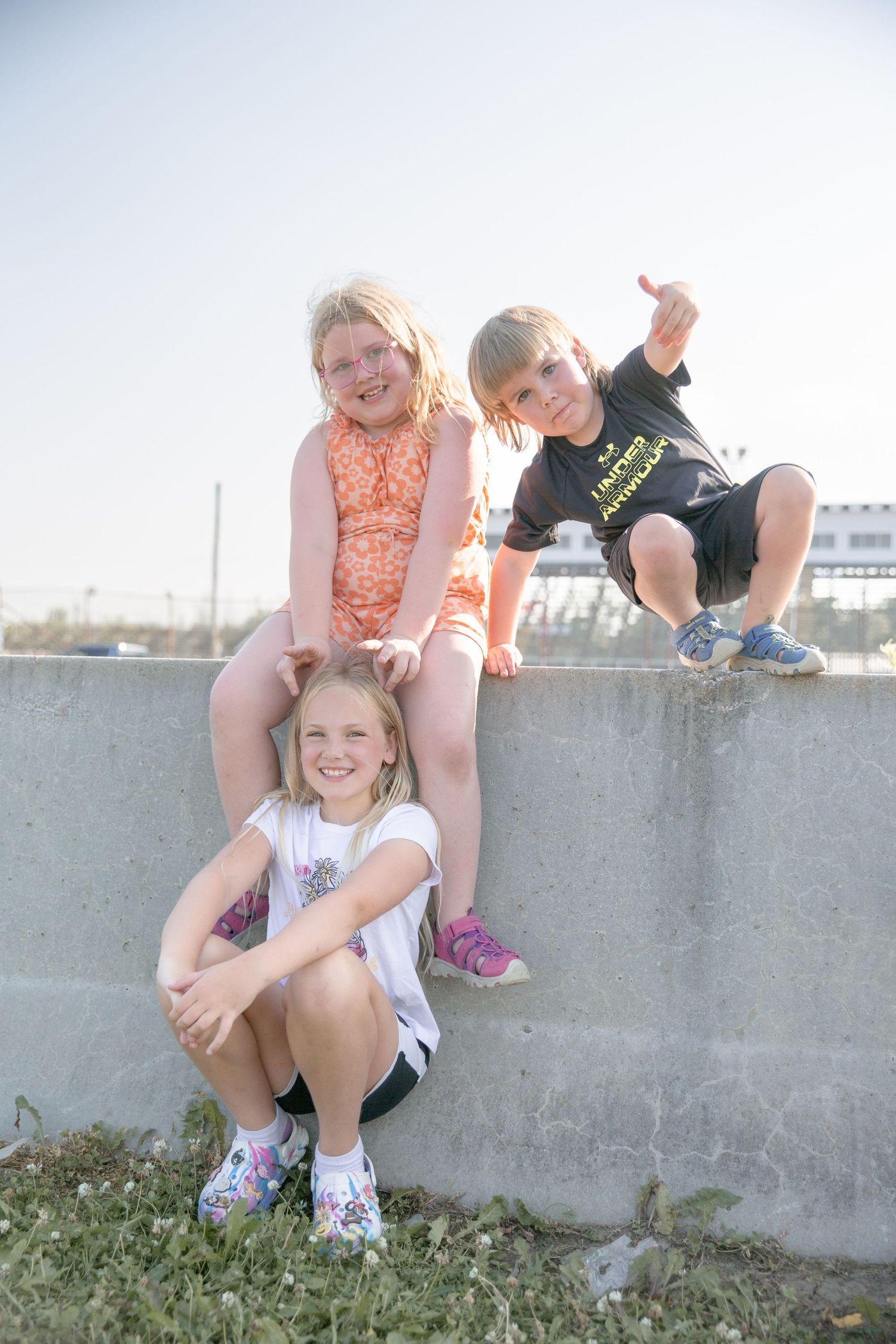 Three young children sit and pose playfully on a concrete wall on a sunny day outdoors.