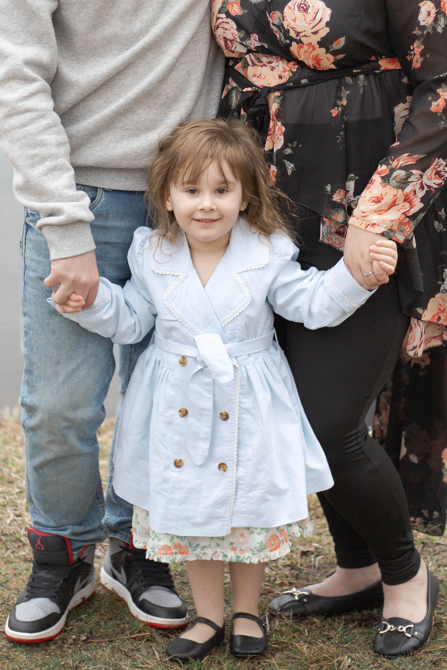 A small child in a light blue coat dress and floral skirt holds hands with adults wearing jeans and a floral dress.