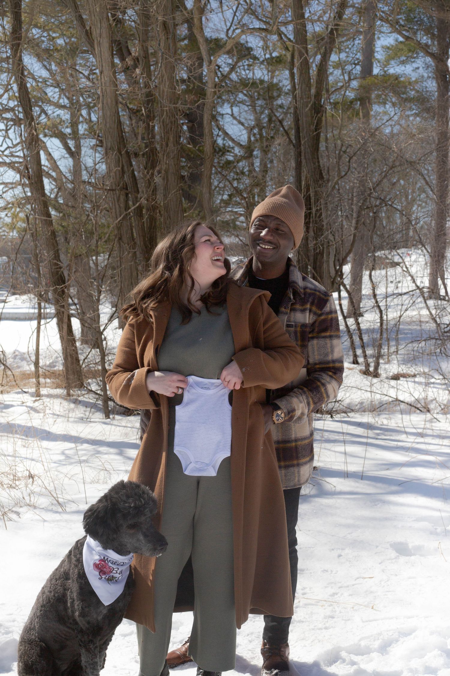 A couple holds a baby onesie while their dog looks on in a snowy winter forest.