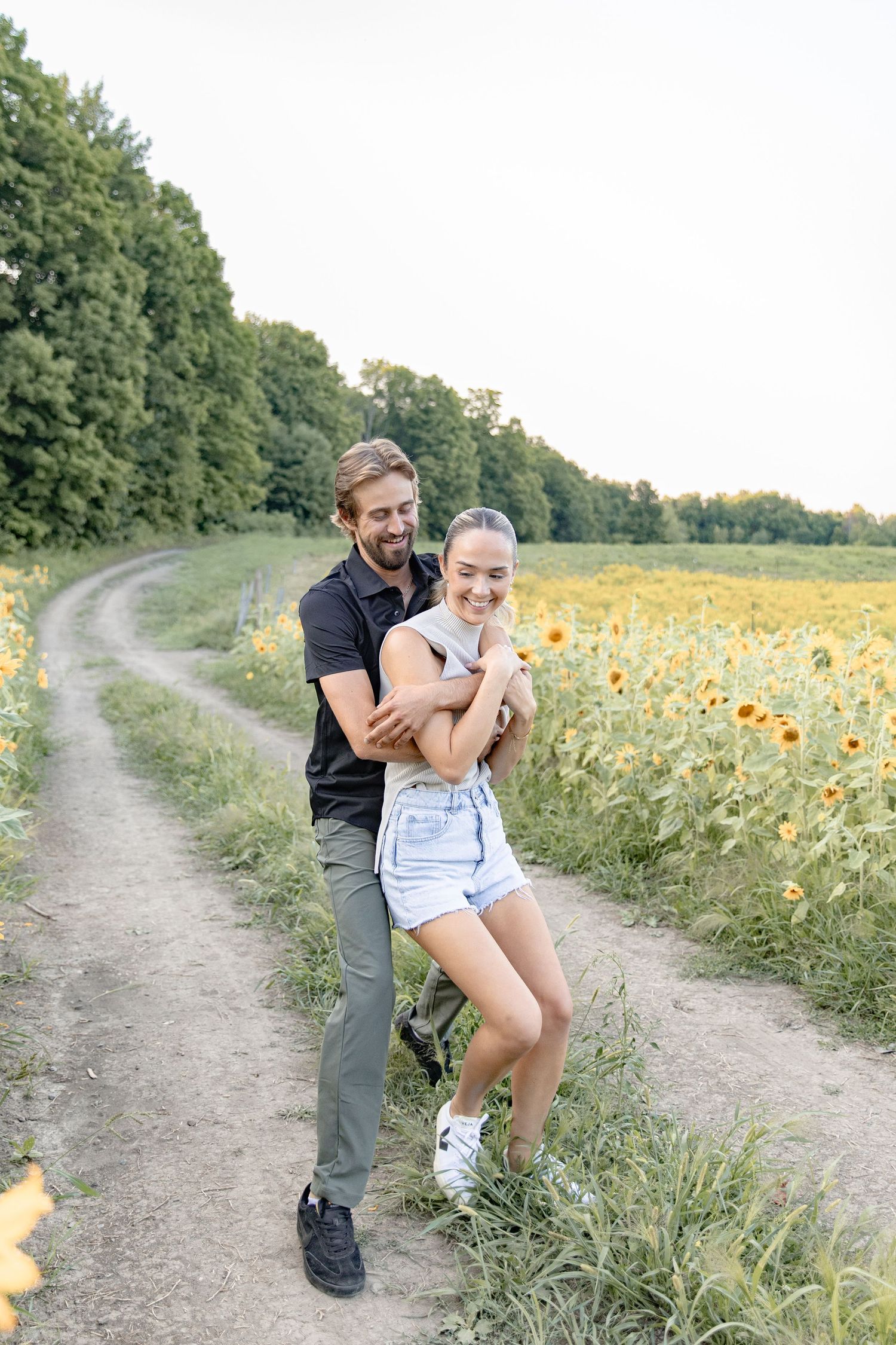 A couple playfully poses along a dirt path next to a vibrant sunflower field on a sunny summer day.