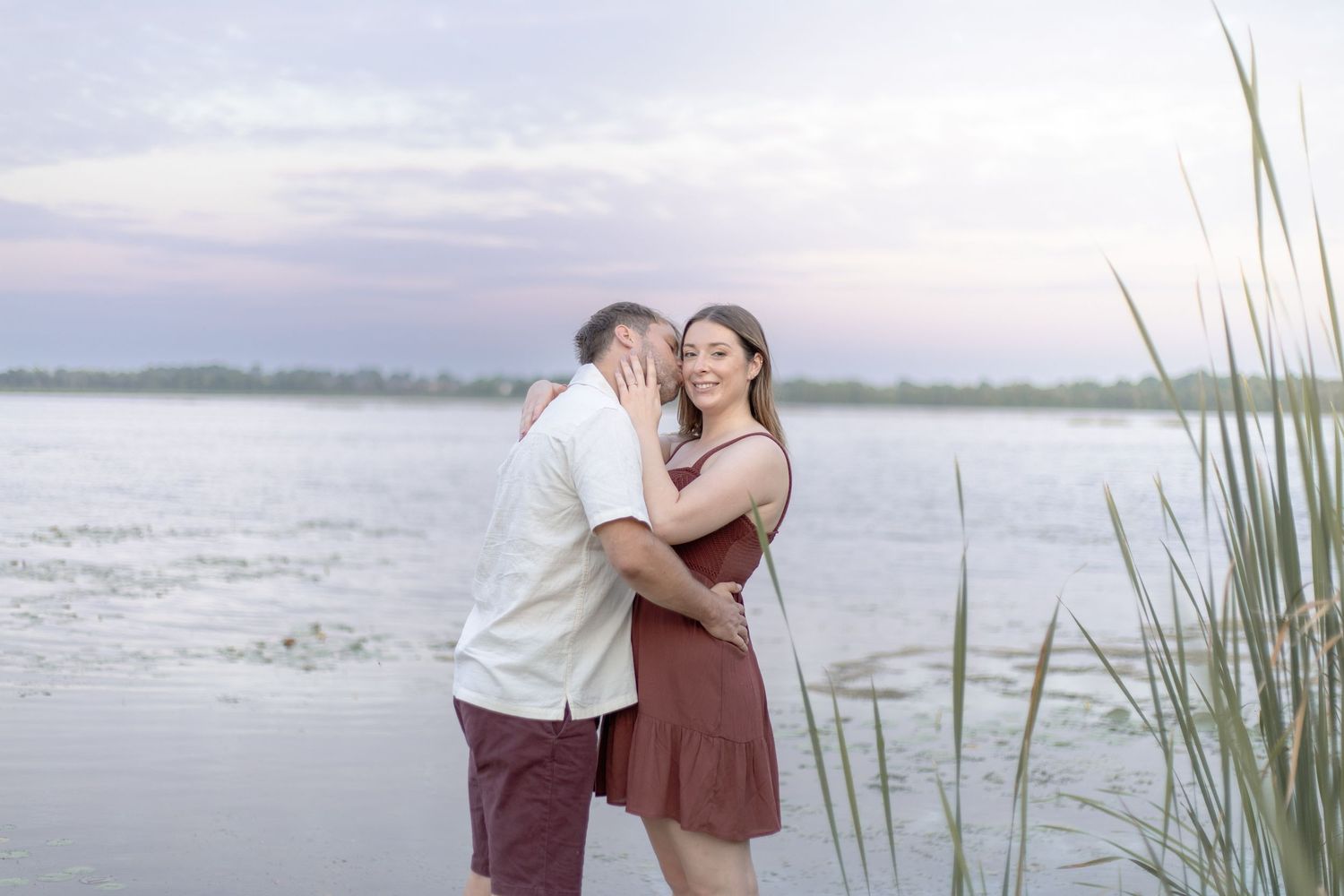 A couple shares an intimate moment by the water with tall beach grass and a serene coastal landscape.