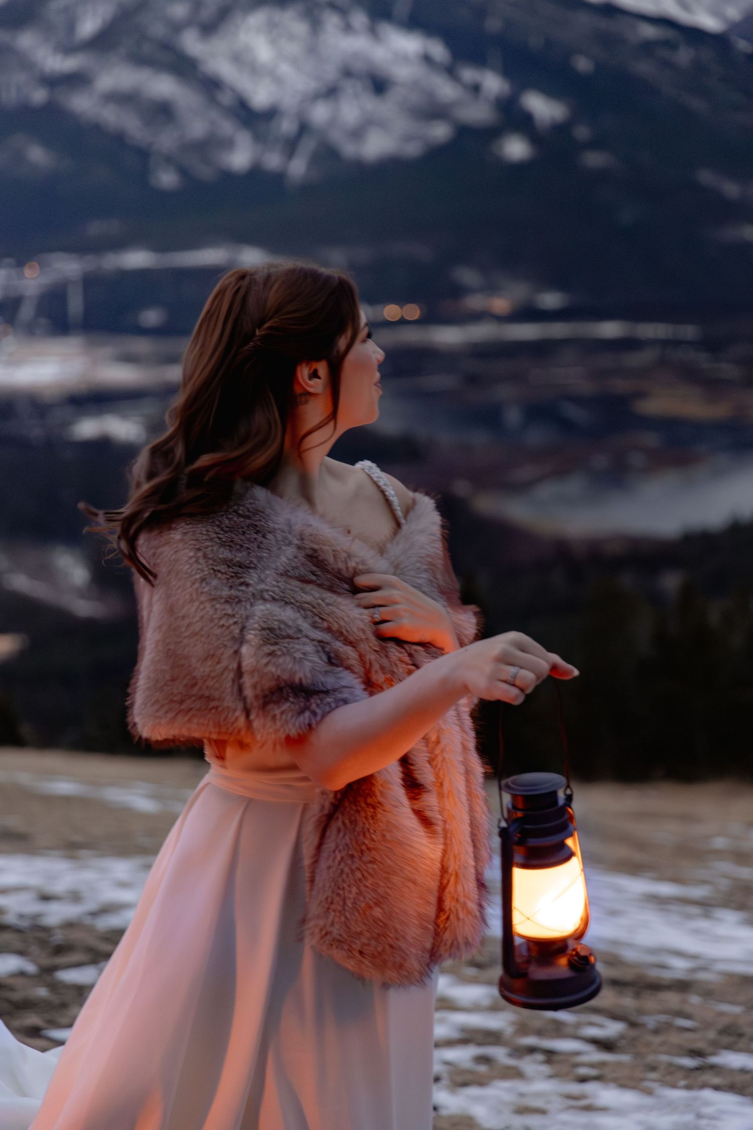 A person in a white dress and fur shawl holds a lantern at dusk in a snowy mountain landscape.