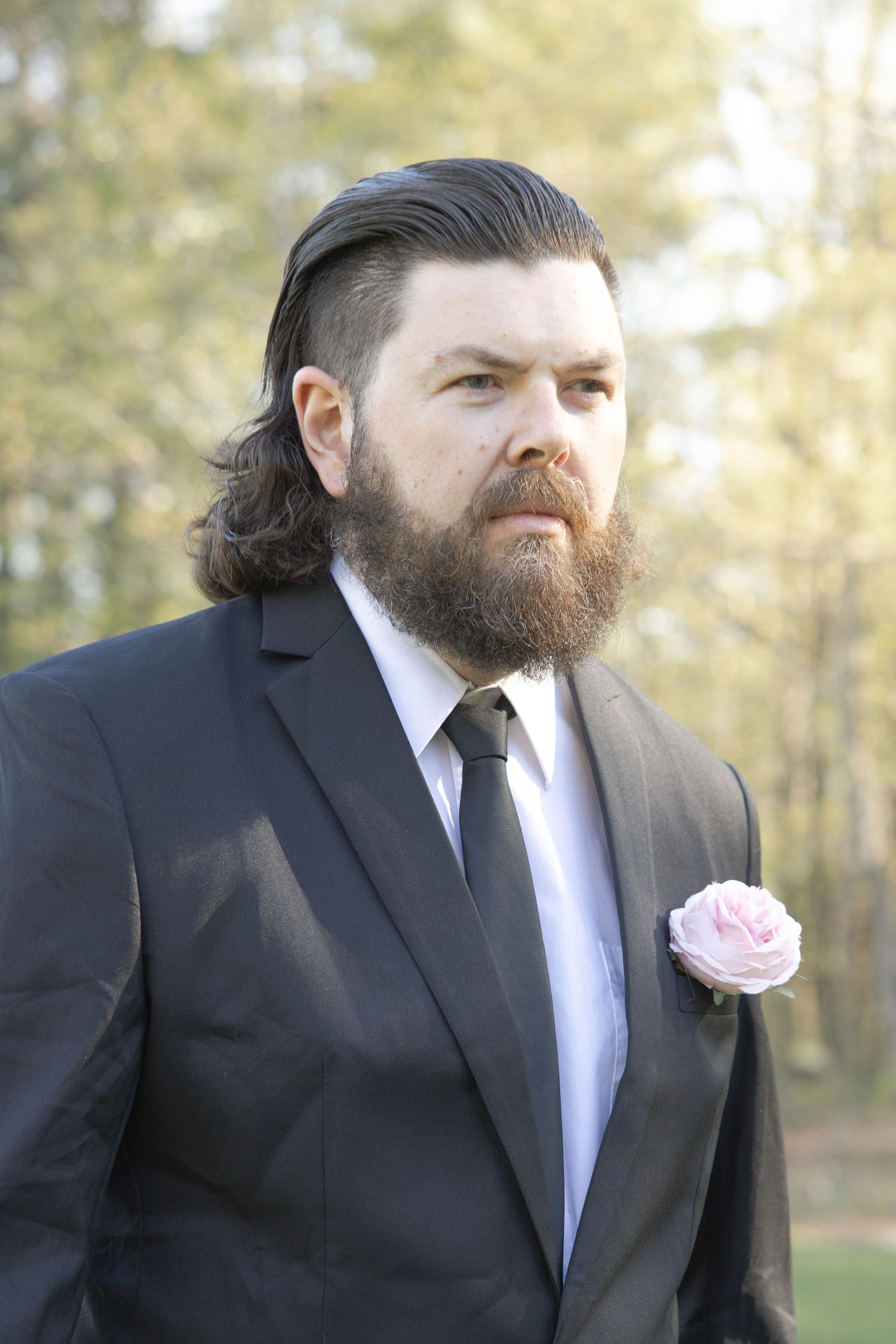 A bearded person in a black suit wearing a pink rose boutonniere poses outdoors on a bright day with trees in background.