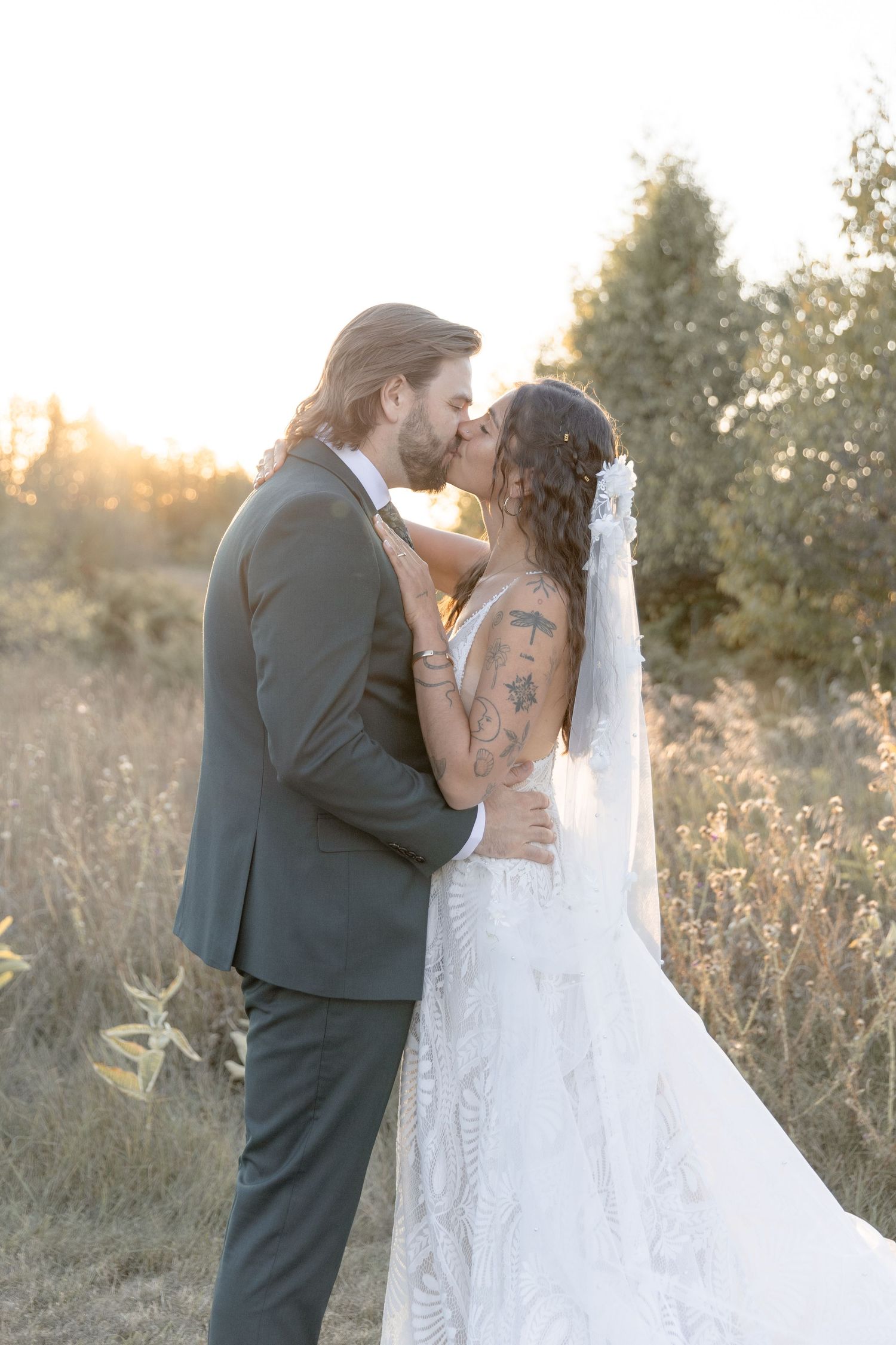 Romantic wedding couple embraces in golden sunlit field with flowing white veil during sunset photoshoot.