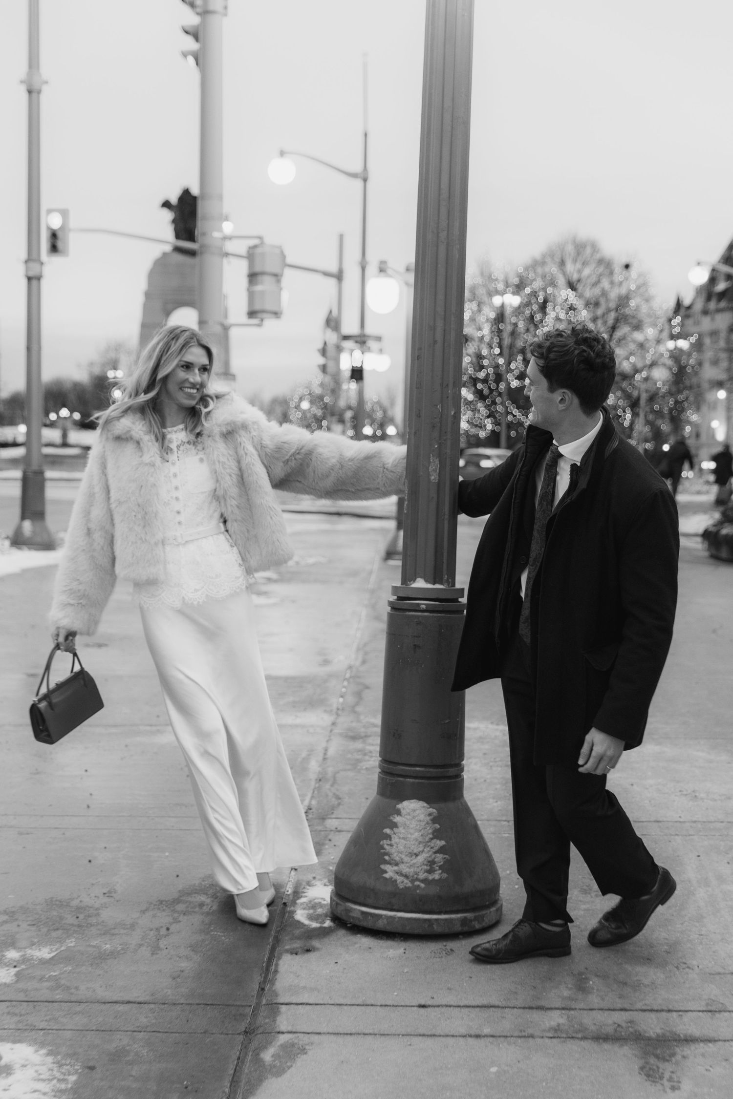 A couple in formal attire poses on a city street corner in black and white, with a vintage lamp post in the background.