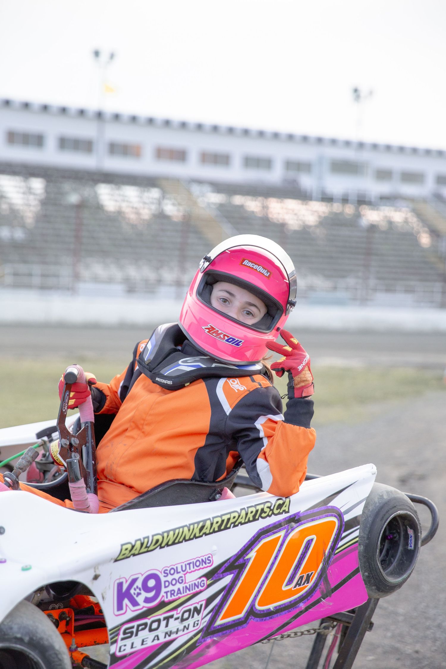 Young go-kart racer in orange racing suit and pink helmet navigates track while piloting white racing kart with K9 sponsorship.