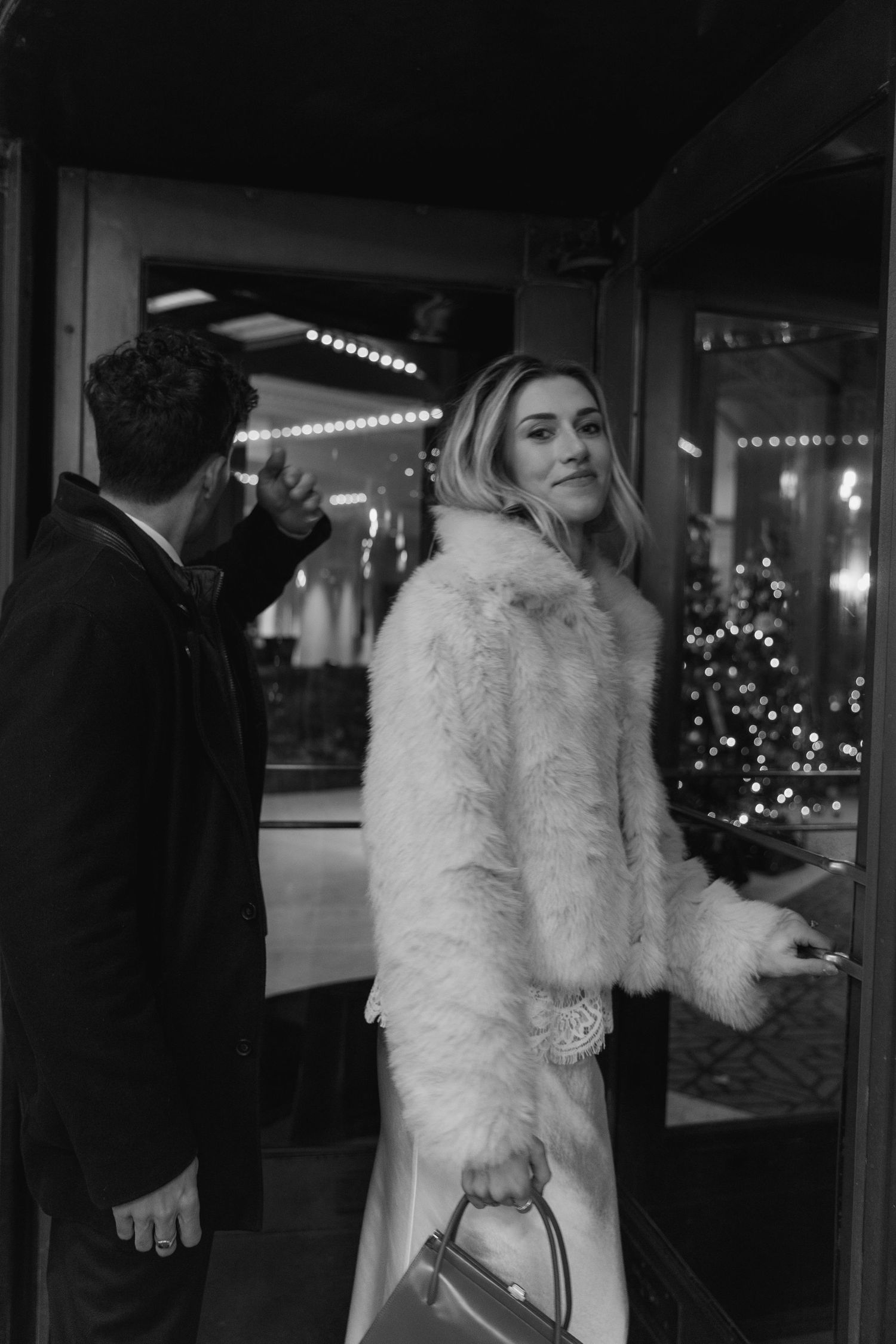 A person in a white fur coat and formal attire stands in a dimly lit venue with Christmas lights visible in background.