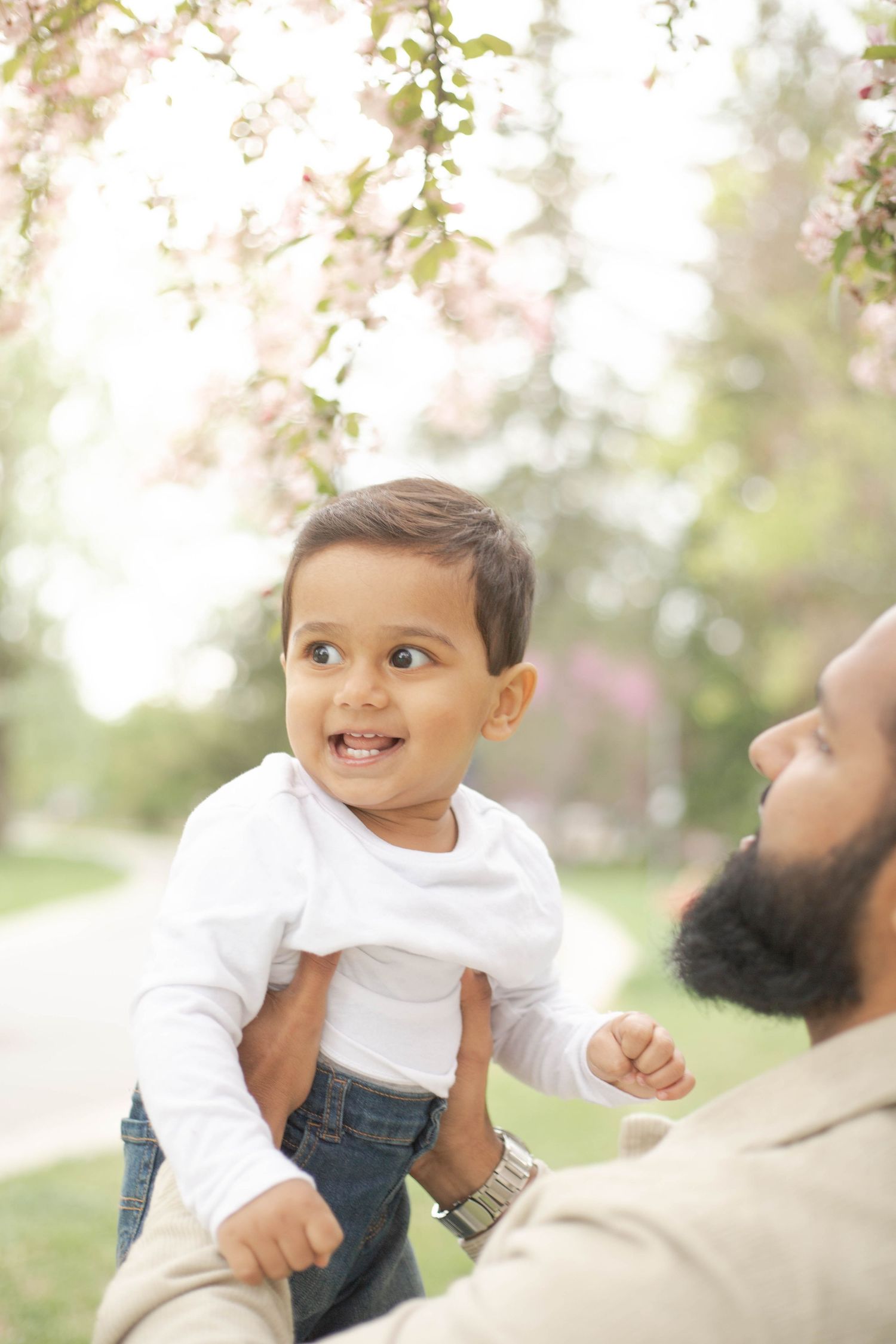 A smiling baby wearing a white shirt and jeans plays outdoors in a sunny garden setting with blurred blossoms.
