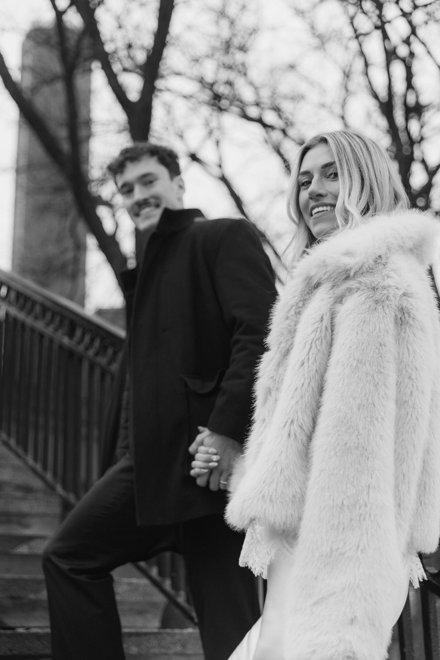 A couple in winter attire poses together on steps during an outdoor photoshoot in black and white.