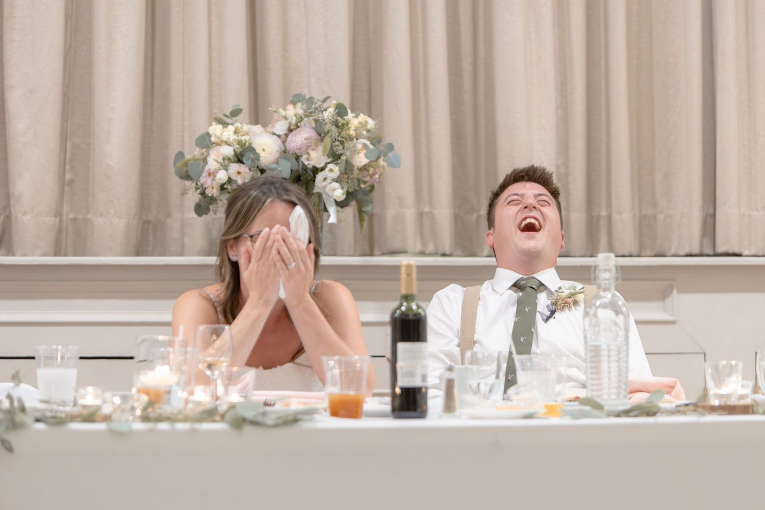 Wedding guests laugh and share joyful moments during reception at tables decorated with floral centerpieces.
