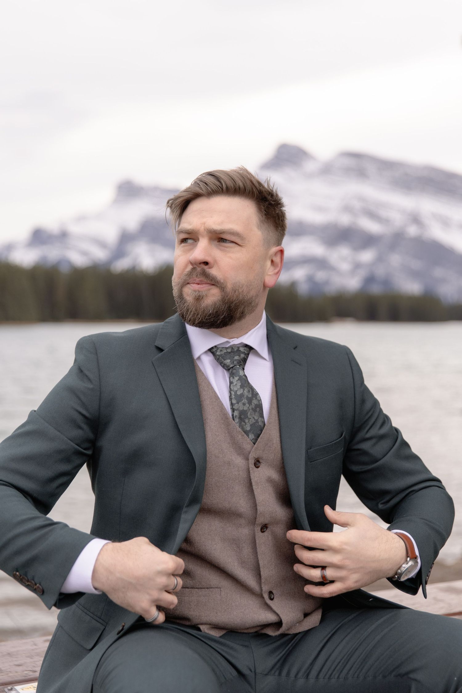 A person in a dark suit and brown vest poses against a snowy mountain landscape by a lake.