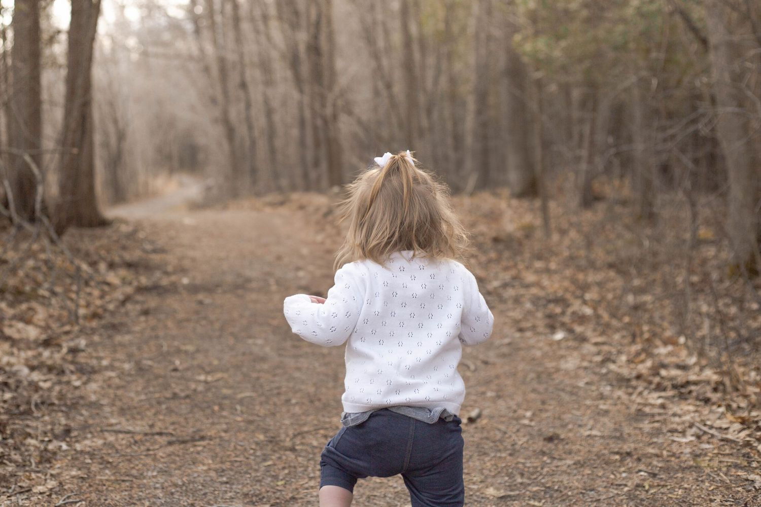 Small child in a white sweater runs down a leaf-covered forest path on an autumn day.