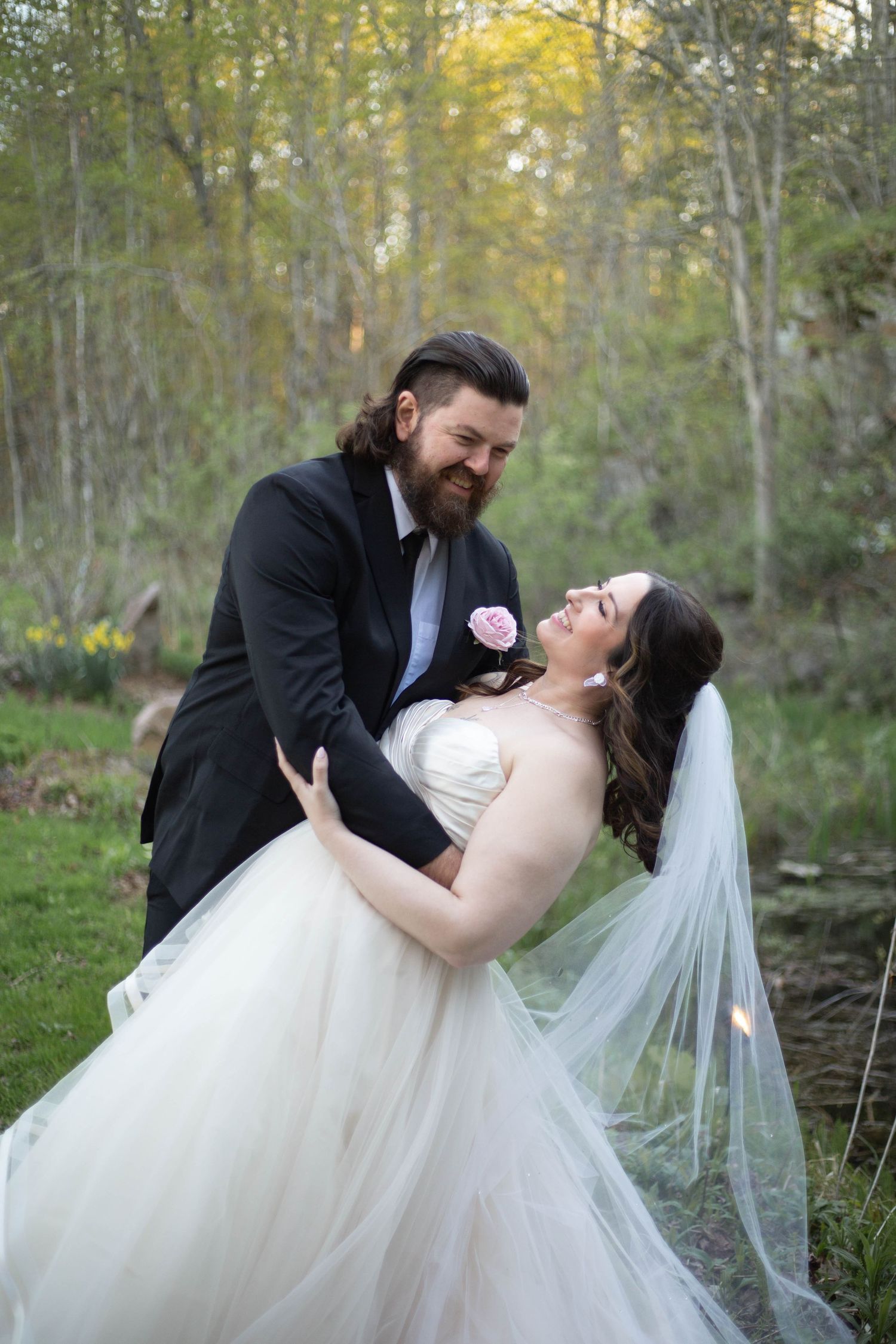 A joyful couple dances outdoors in wedding attire, dipping and laughing together in a wooded setting.