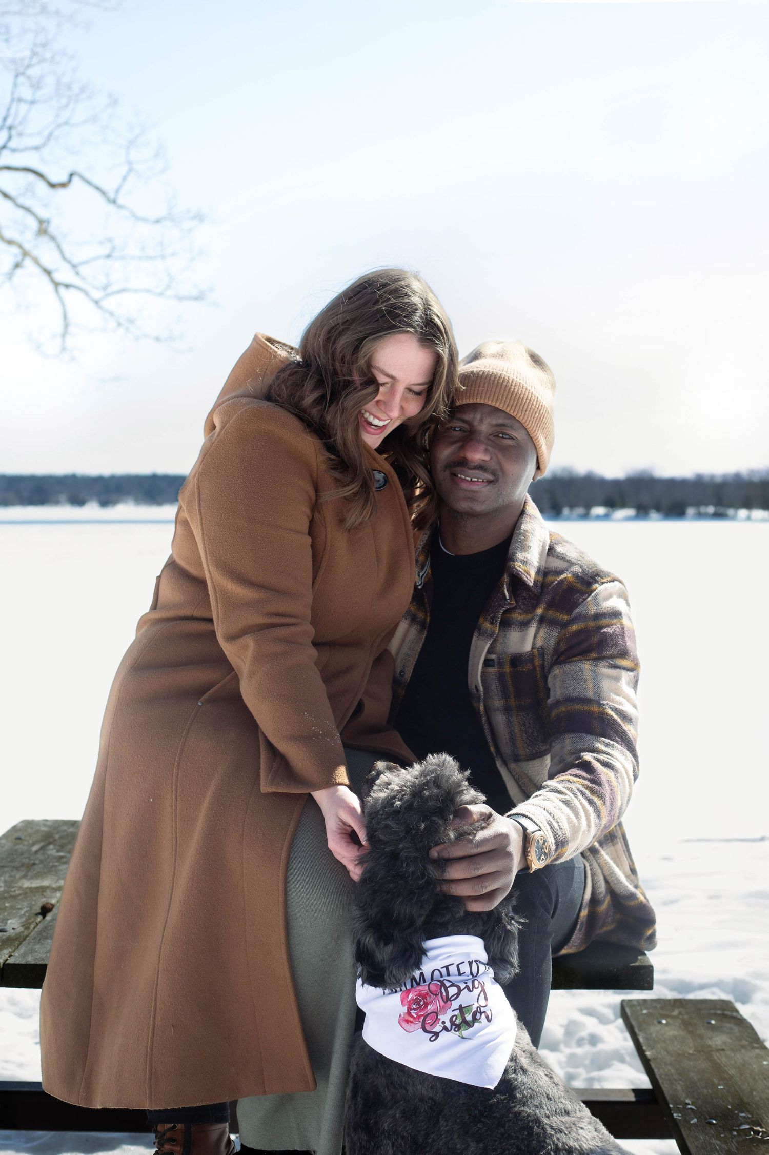 A joyful couple poses with their dog on a winter dock by a frozen lake. Dog wears 