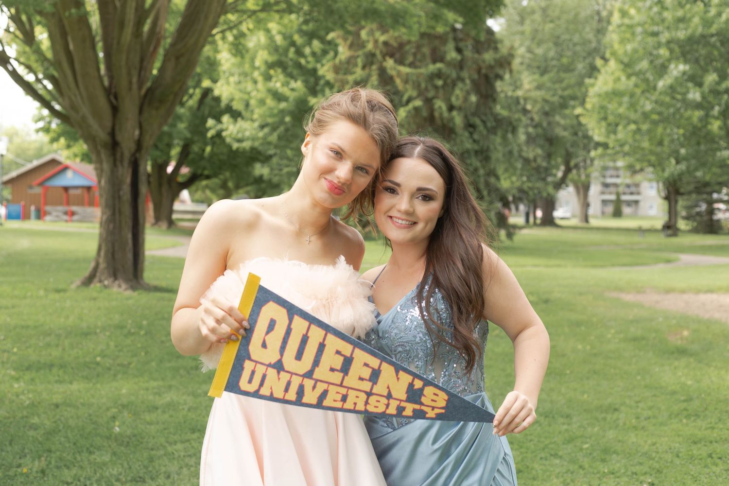 Two friends pose together outdoors on a sunny day while holding a Queens University pennant flag in a park setting.