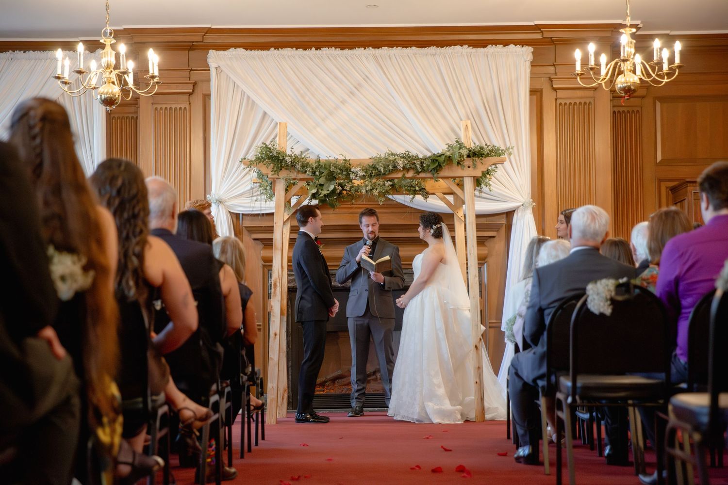 A wedding ceremony takes place in an elegant wood-paneled room with chandeliers and draped white fabric decorations.